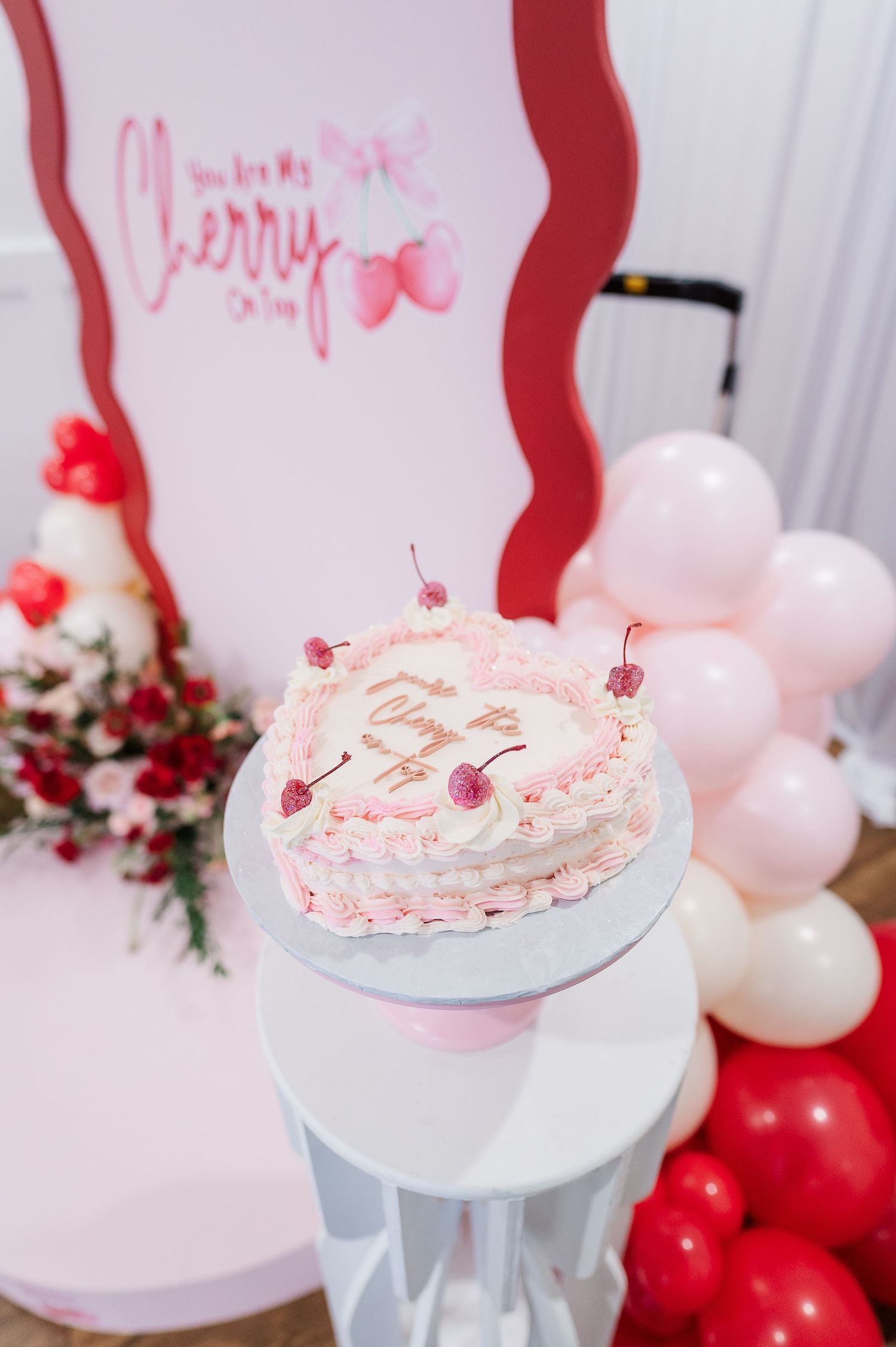 Pink and white crepe cake decorated with cherries on a pedestal stand surrounded by balloons.