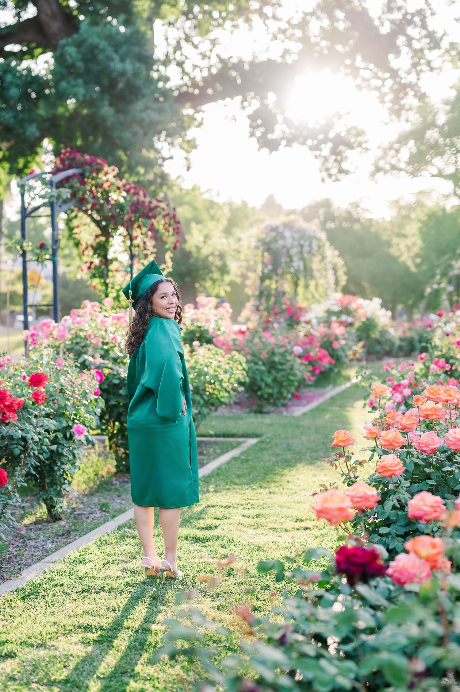A woman in emerald green stands among blooming rose gardens on a sunny day.