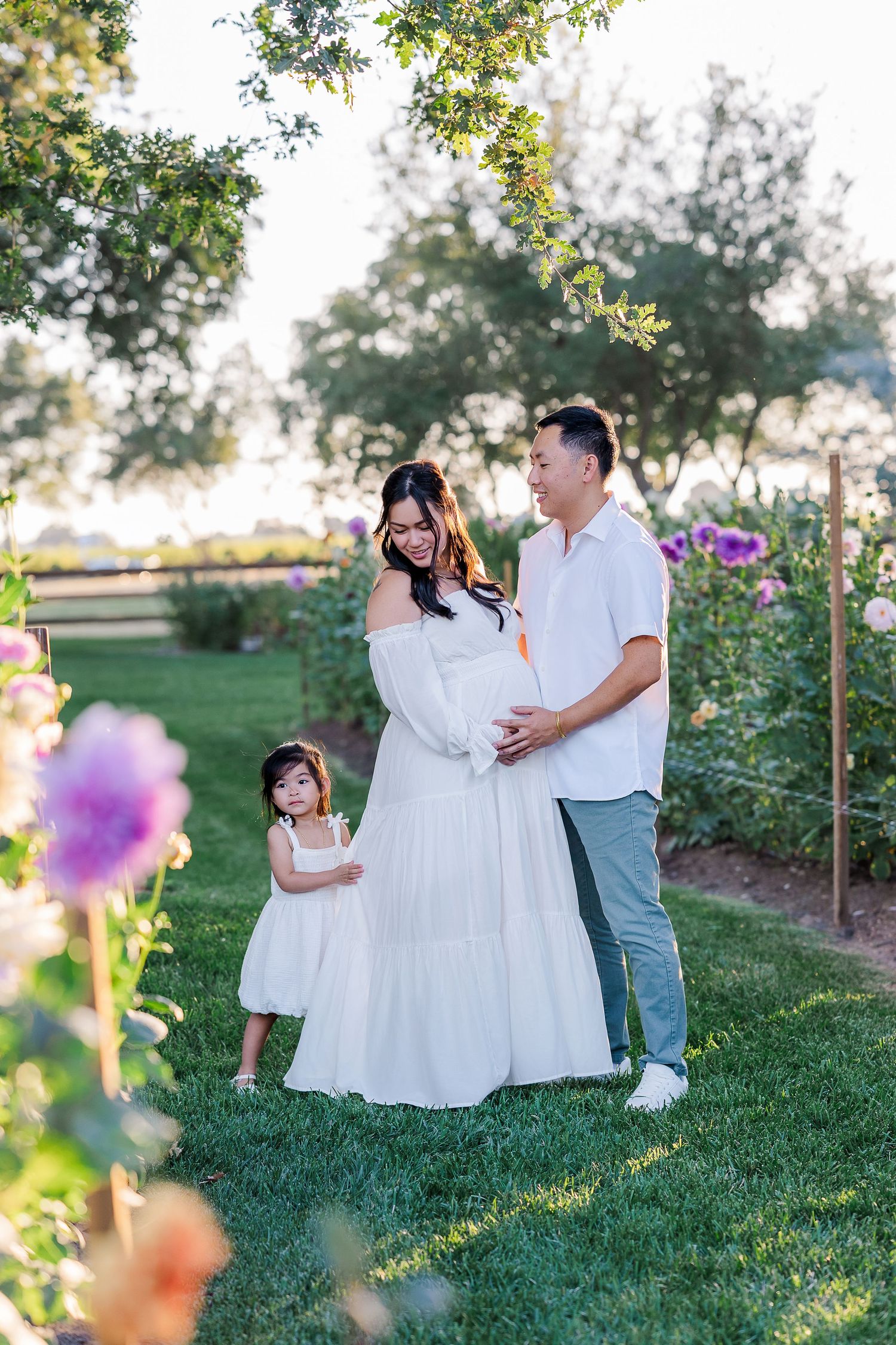 Pregnant woman in white dress with husband and daughter in a flower garden during golden hour maternity photo session.