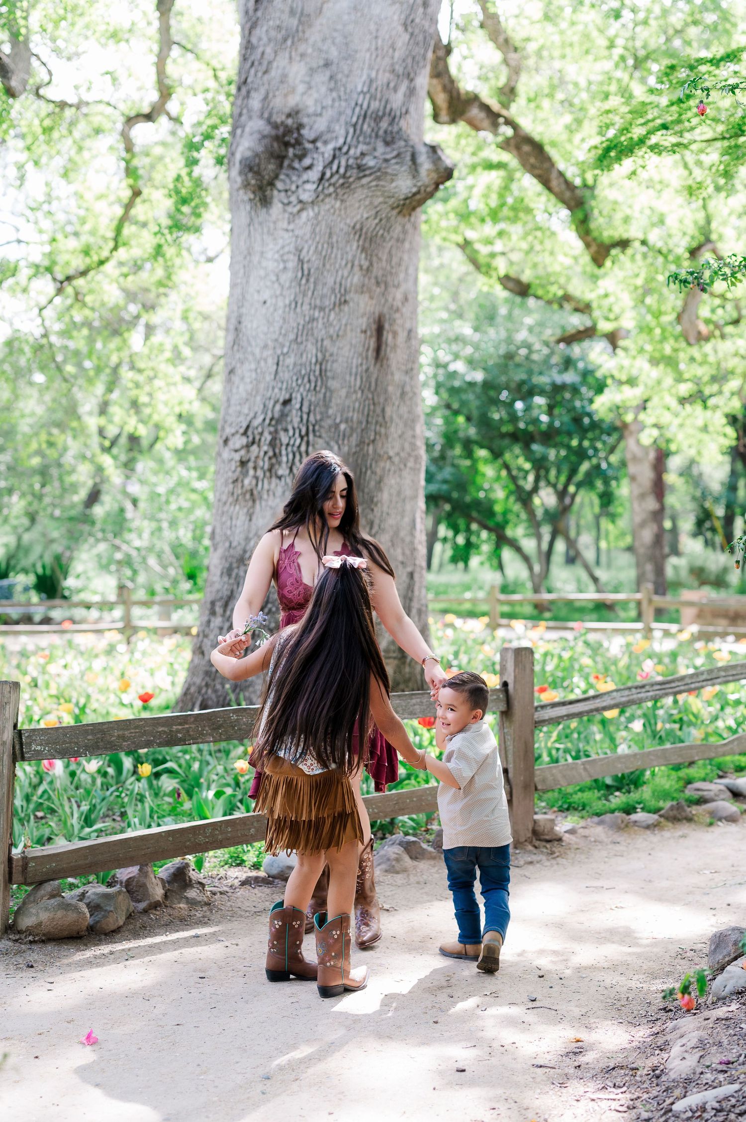 Two people in western wear walking with a small child along a garden path near a large tree.