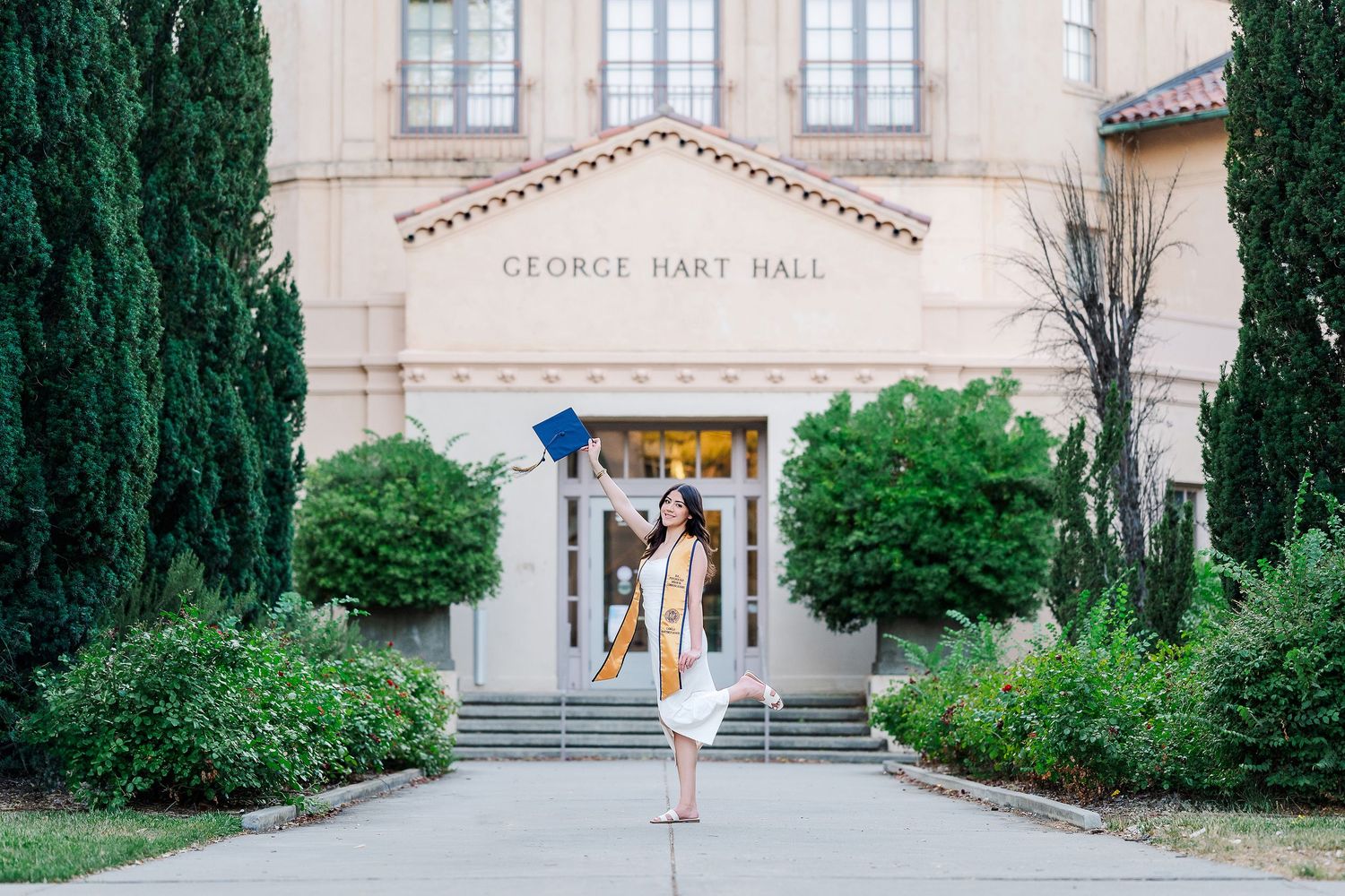 A graduate stands in front of George Hart Hall building entrance flanked by cypress trees on a sunny day.