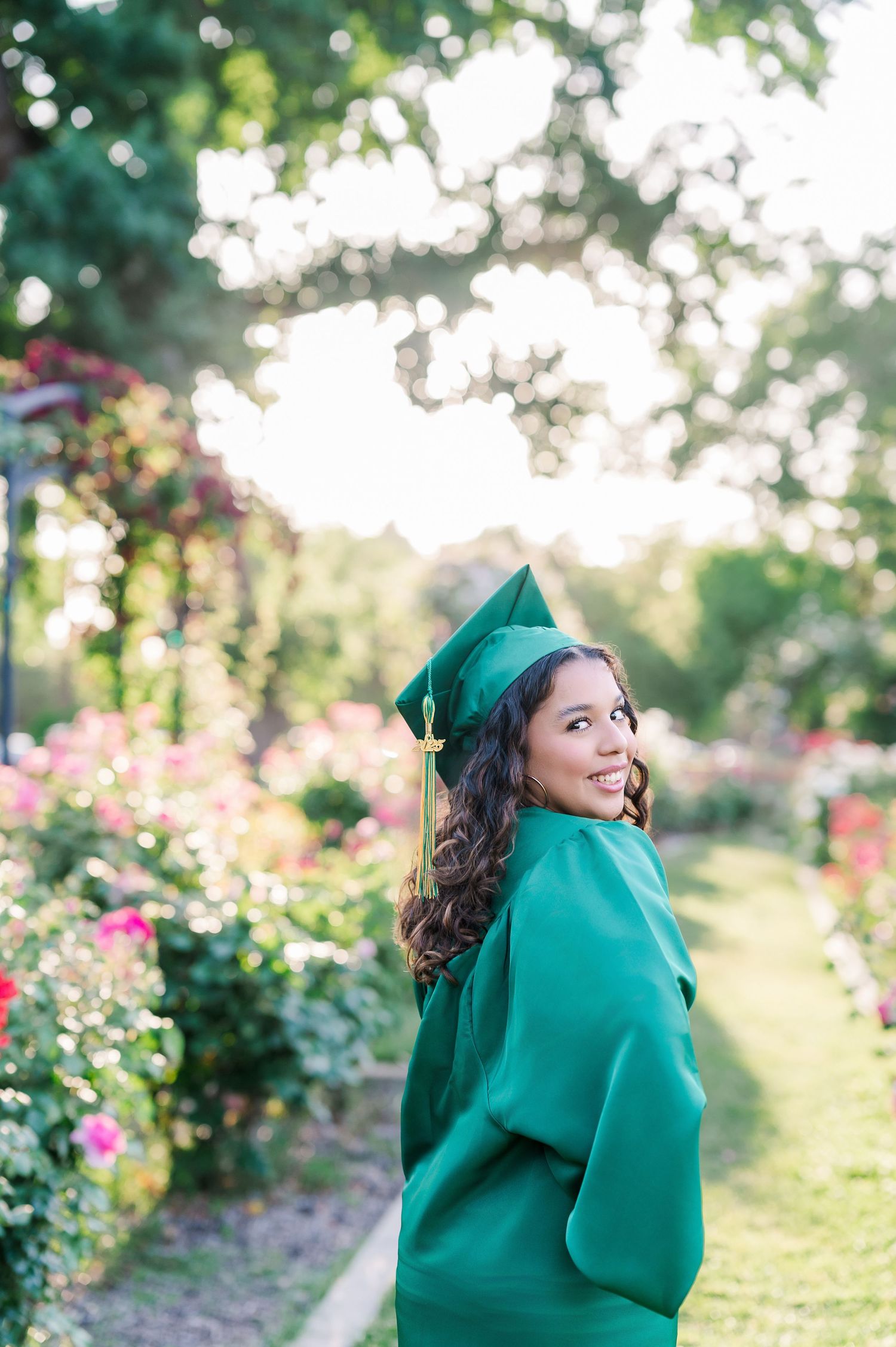 Smiling graduate in green cap and gown posing in a lush garden with colorful flowers and sunlight.
