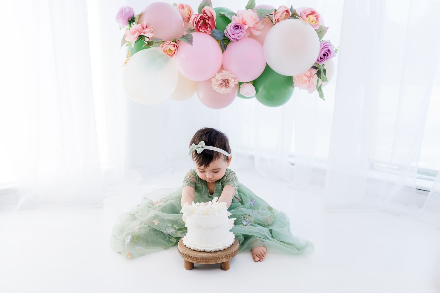Baby girl in green tutu dress sits before white smash cake under pink balloon arch with flowers.