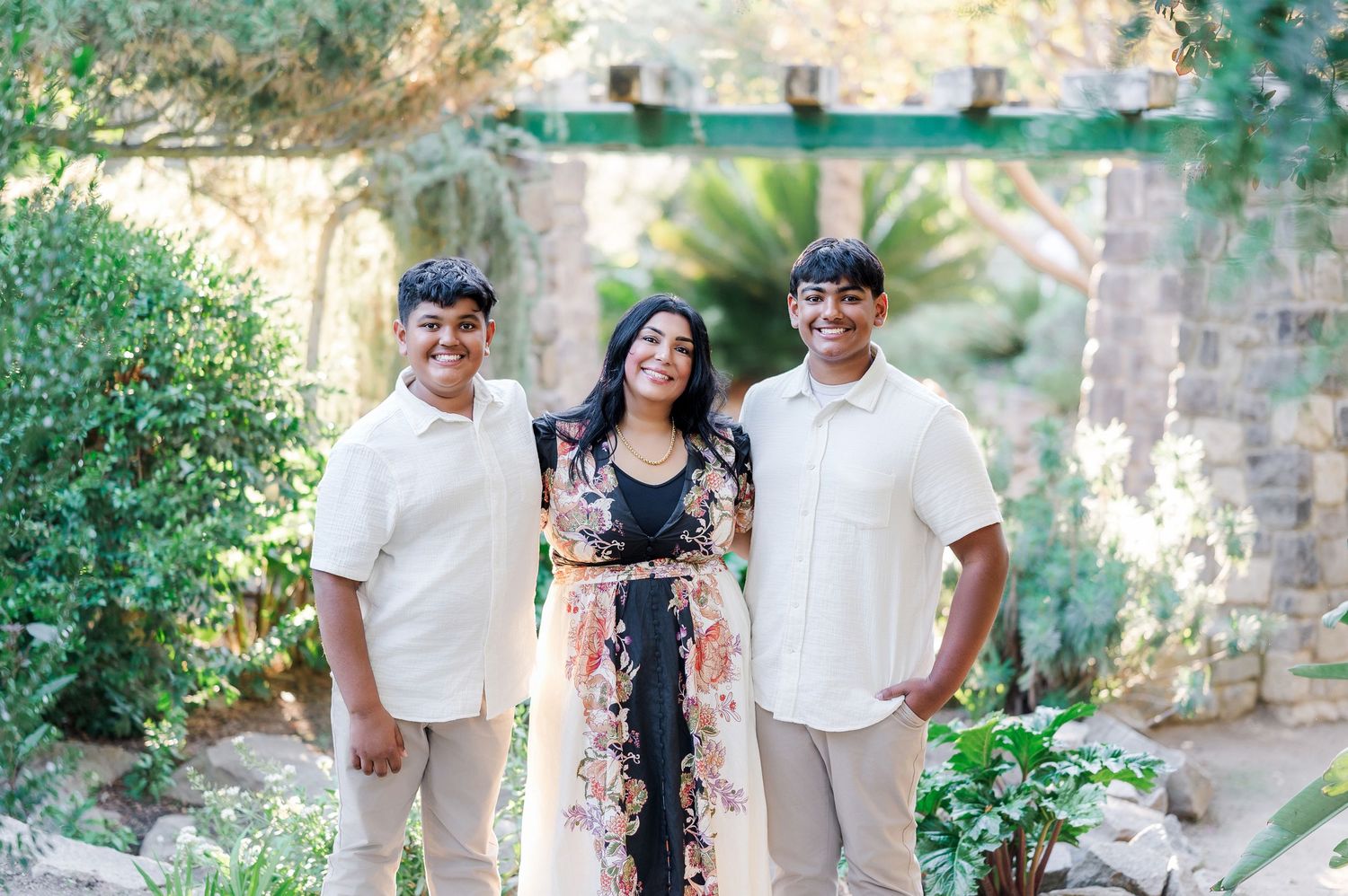 A family poses together outdoors in a garden setting with stone walls and tropical greenery.
