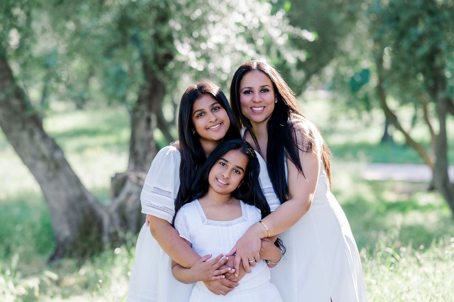 Three people wearing white dresses pose together outdoors in a sunlit garden setting with trees in the background.