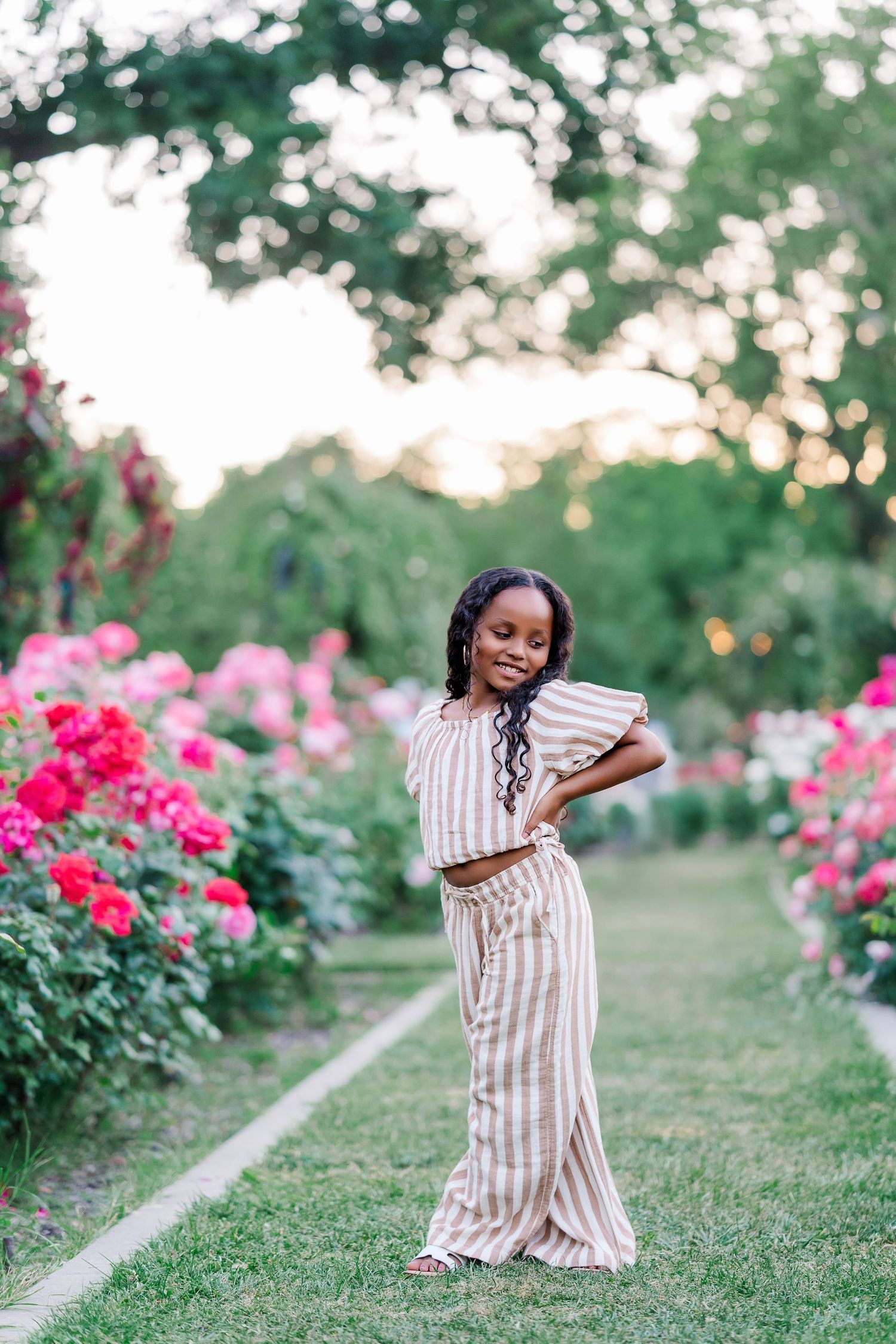 Person in striped outfit poses elegantly among pink rose garden at golden hour.