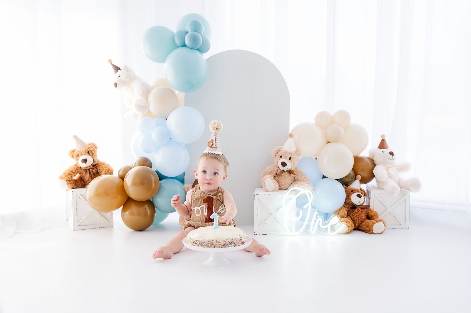 Baby's first birthday cake smash with teddy bears, blue and gold balloons, and white backdrop.