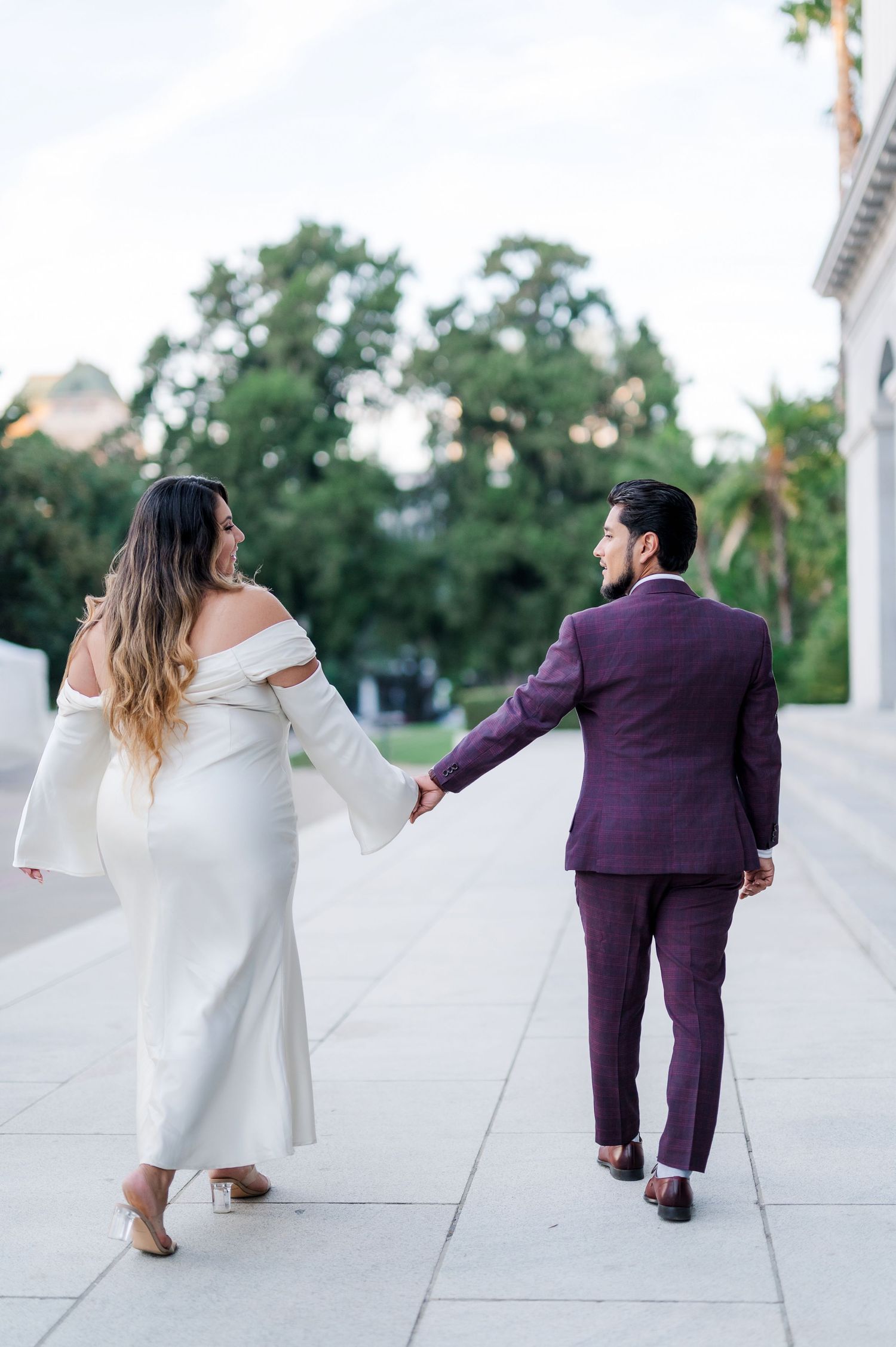 Couple walking hand in hand, woman in white off-shoulder dress, man in purple suit, outdoor elegant setting.