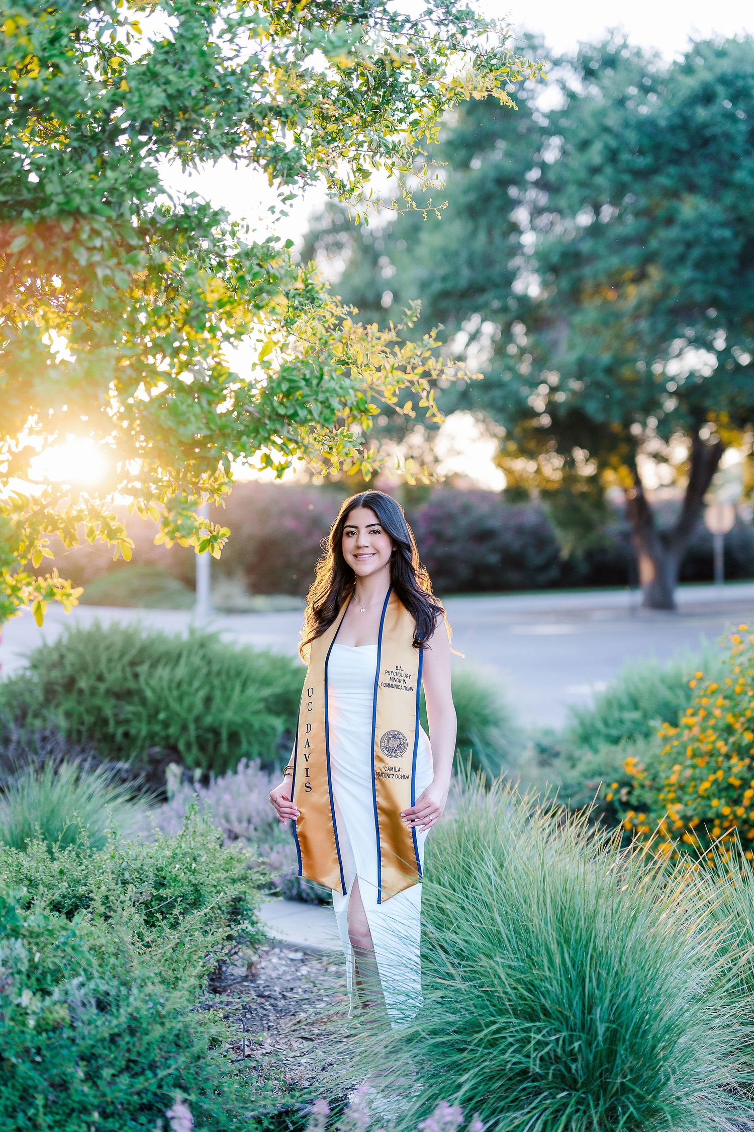 Graduate posing in a garden setting with golden sunlight filtering through trees and ornamental grasses.