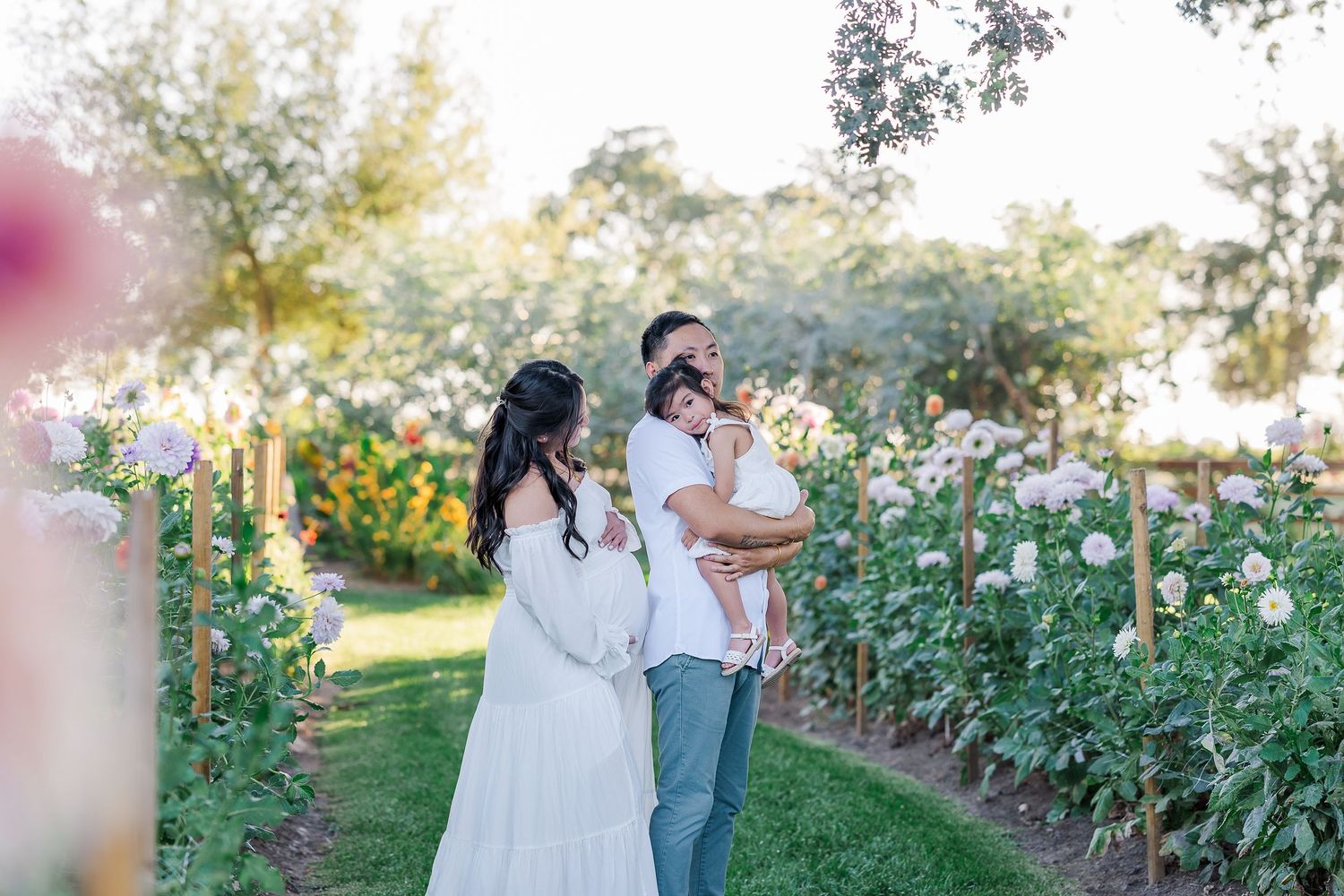 Family of three posing in a lush rose garden, mother in white dress, father holding baby, soft natural light. Sacramento Maternity