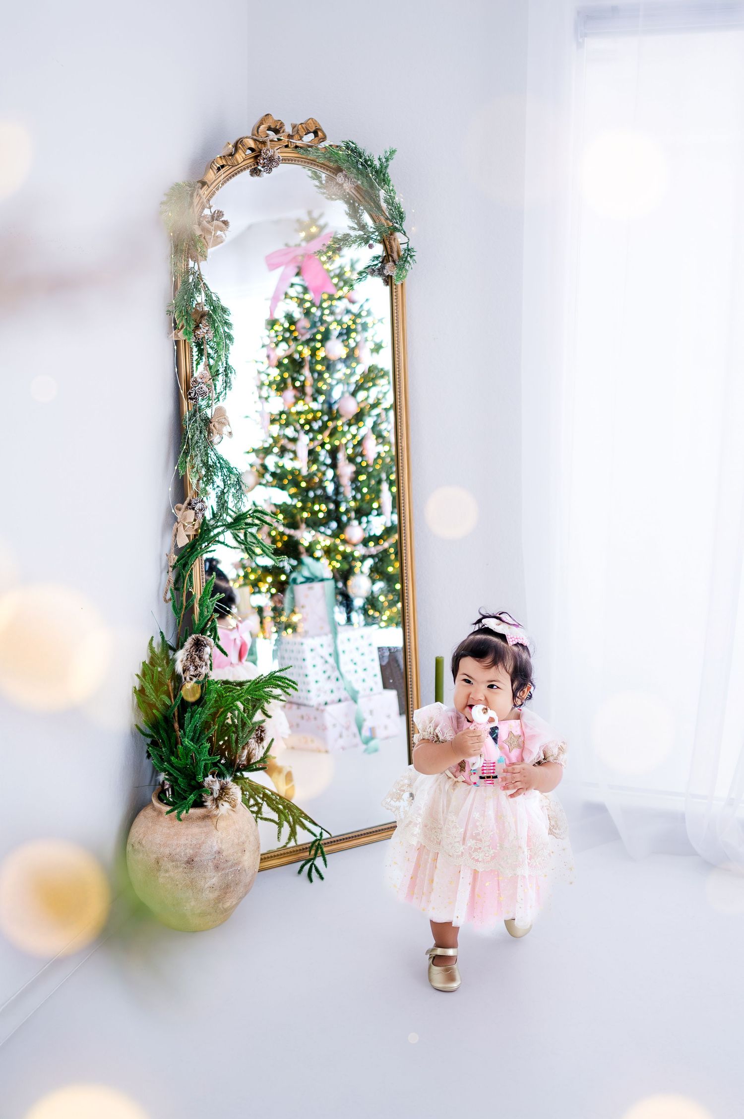 Toddler girl in pink dress stands before ornate gold mirror reflecting Christmas tree in bright white room.