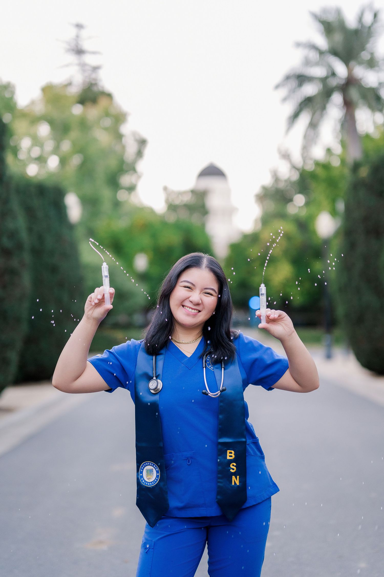 Healthcare worker in blue scrubs celebrates joyfully outdoors with palm trees in background.