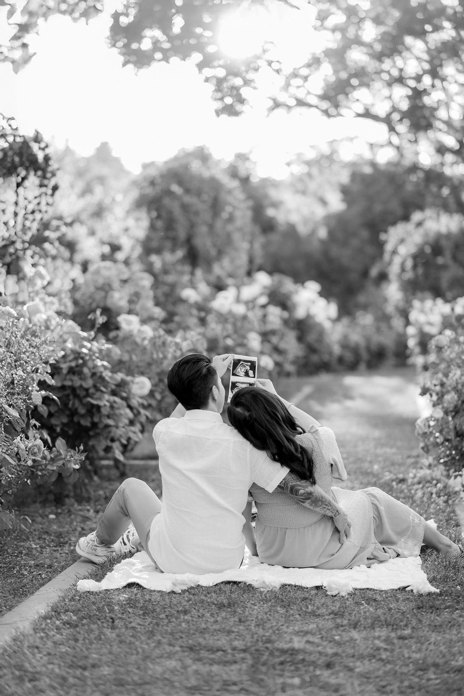 A couple sits on a picnic blanket in a garden while taking a selfie together on a sunny afternoon.