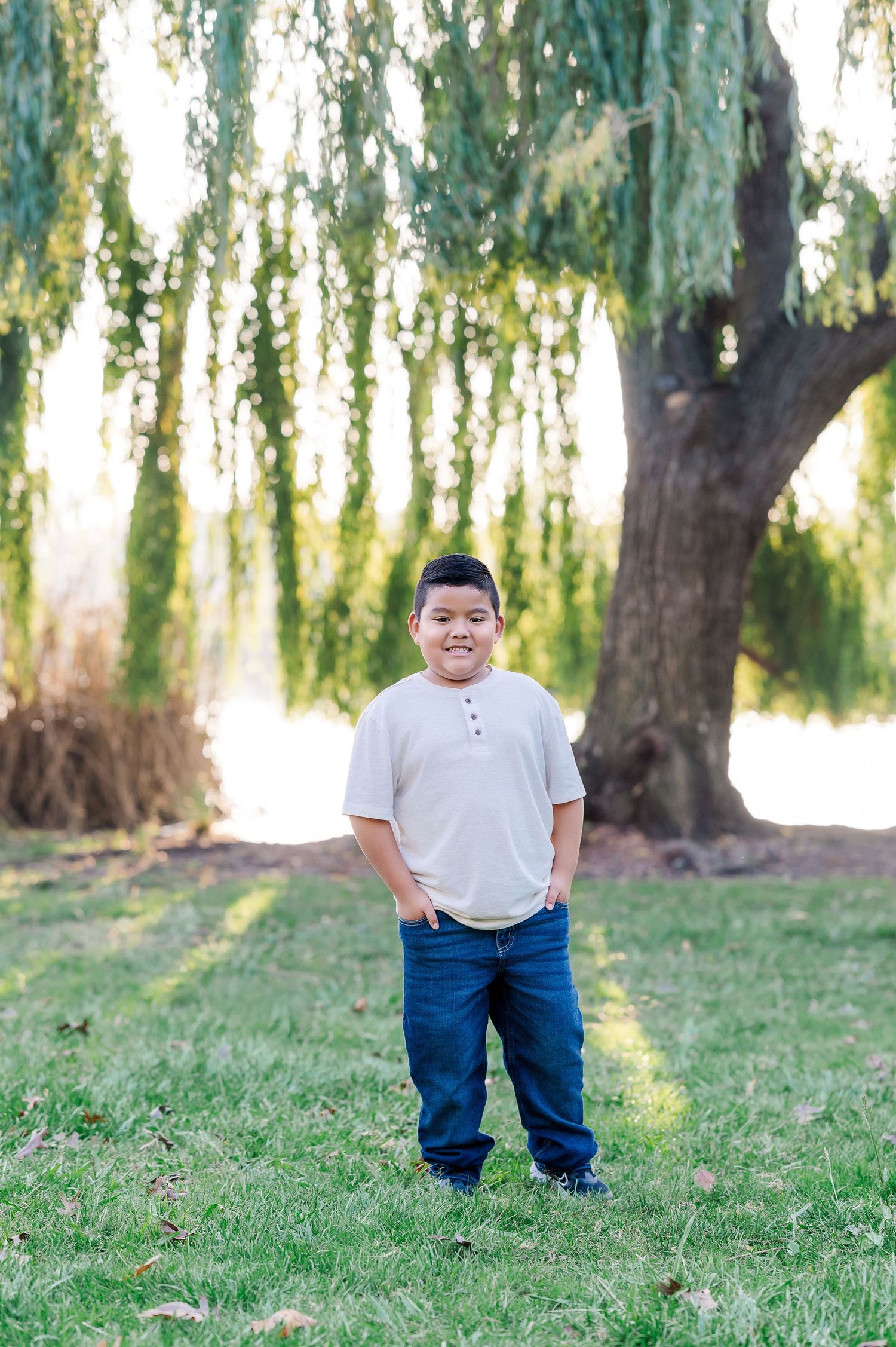 A child stands in a grassy field beneath drooping willow tree branches on a sunny day.
