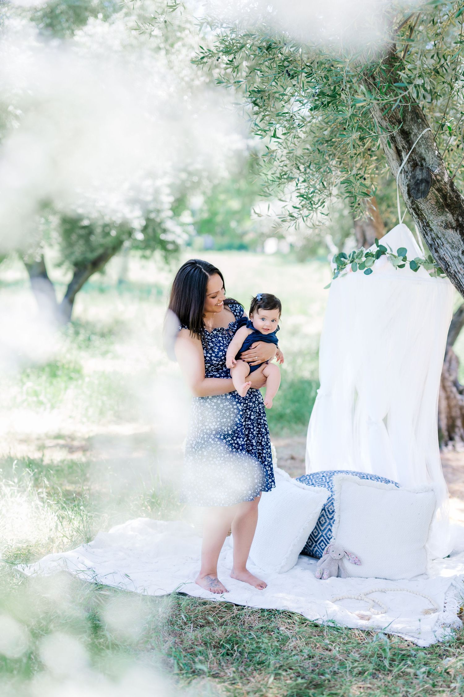 A family shares tender moments together on a white blanket under olive trees in a dreamy outdoor setting.