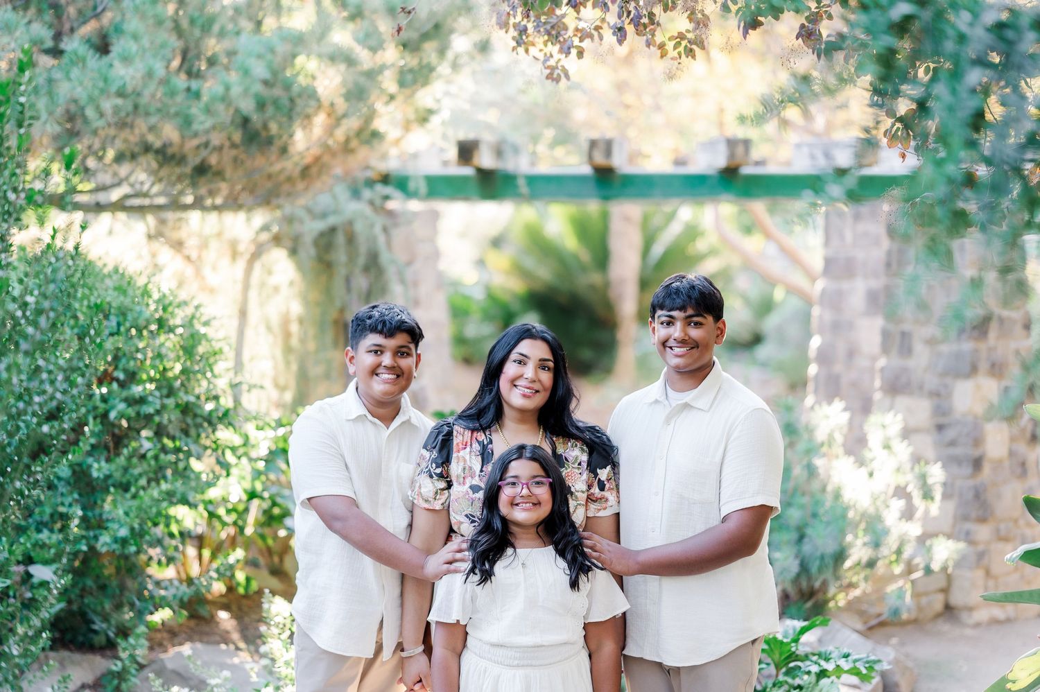 A family wearing coordinated white and beige outfits poses together in a garden setting with stone archway and greenery.
