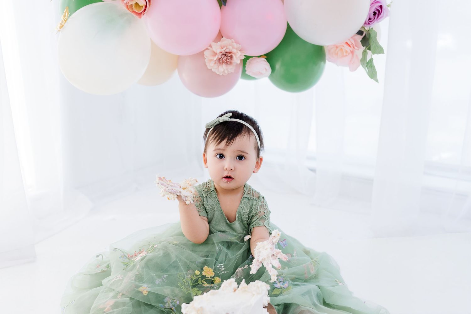 Baby girl in green tulle dress sits under pastel balloon arch with flowers for first birthday cake smash photo.