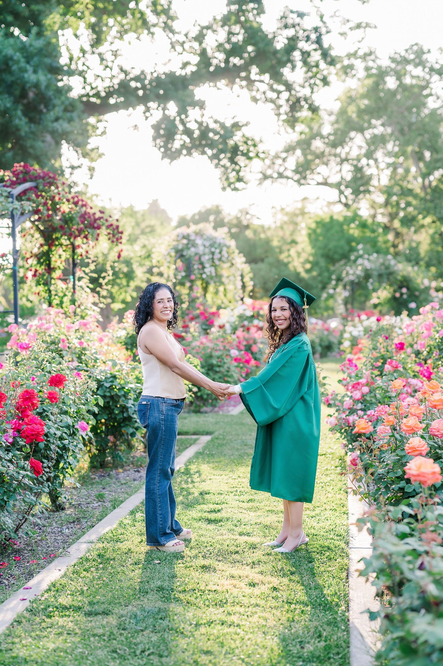 Two women hold hands in a rose garden, one wearing a green graduation cap and gown, celebrating a milestone achievement.