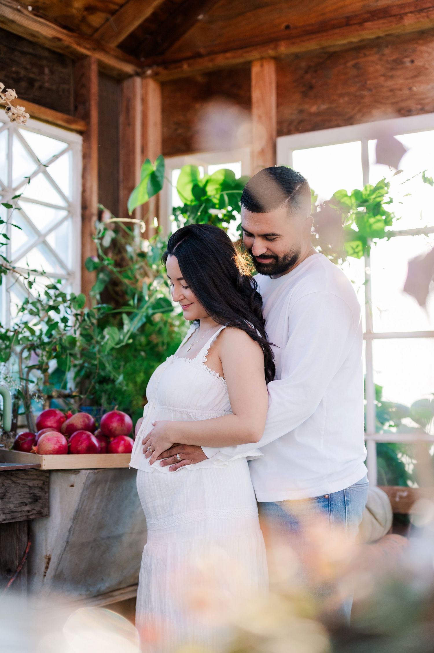 Pregnant couple embracing tenderly in a rustic greenhouse with lush plants and red apples, wearing white outfits.