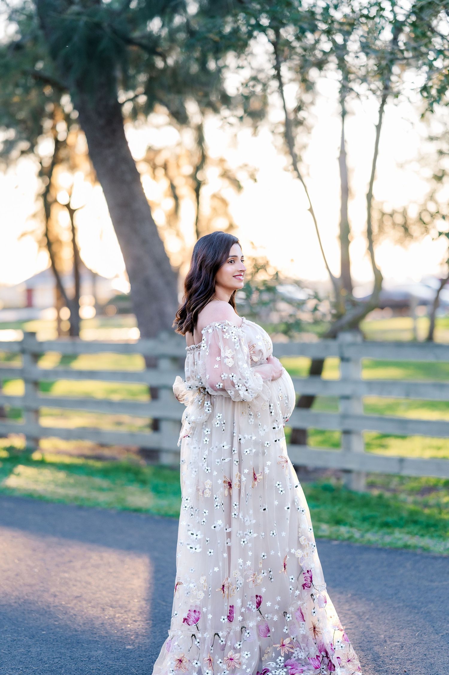 Woman in flowing white floral dress poses for maternity photos on path with wooden fence and trees.