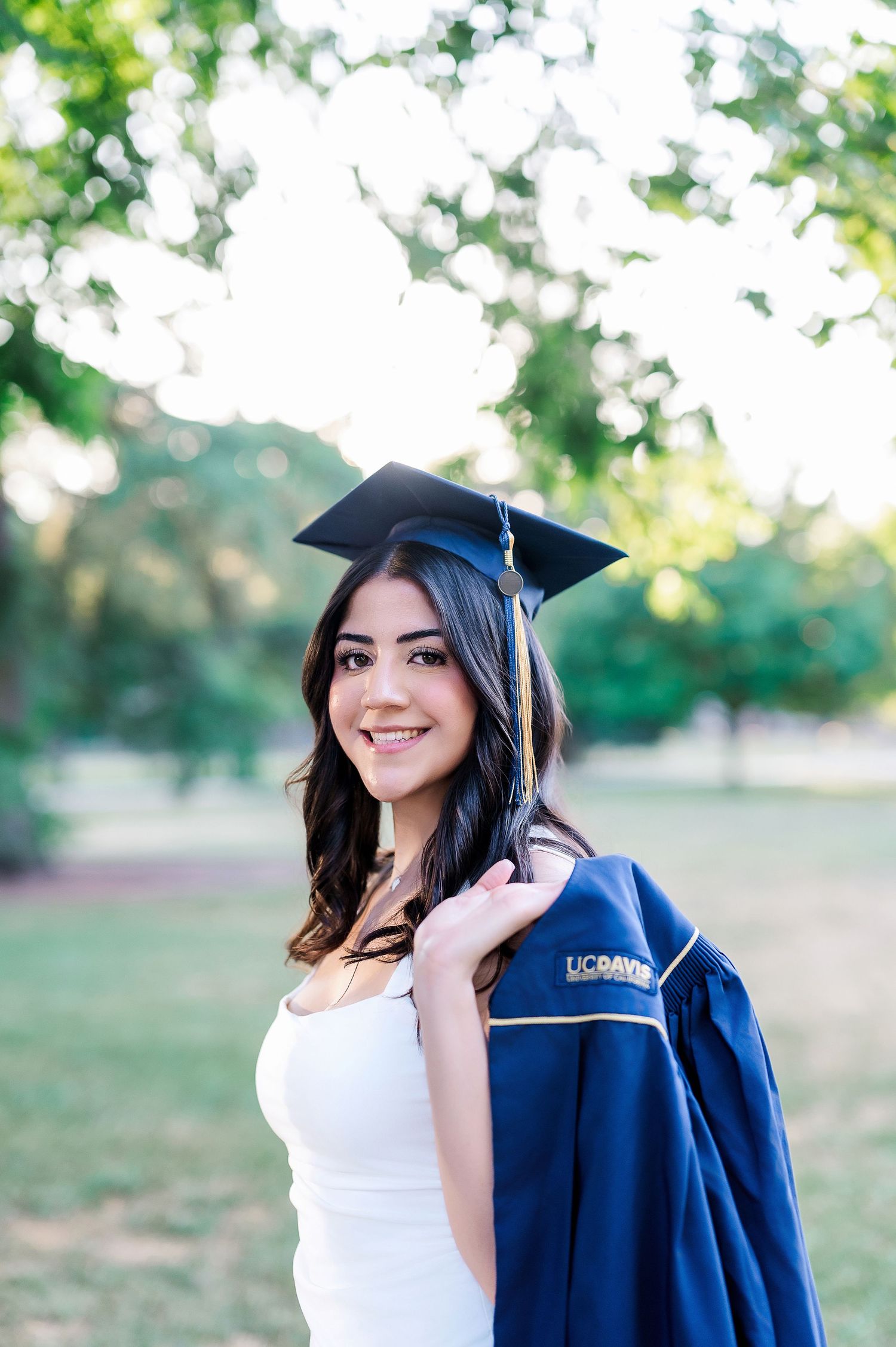 Graduate wearing blue cap and gown poses outdoors in natural sunlit setting.