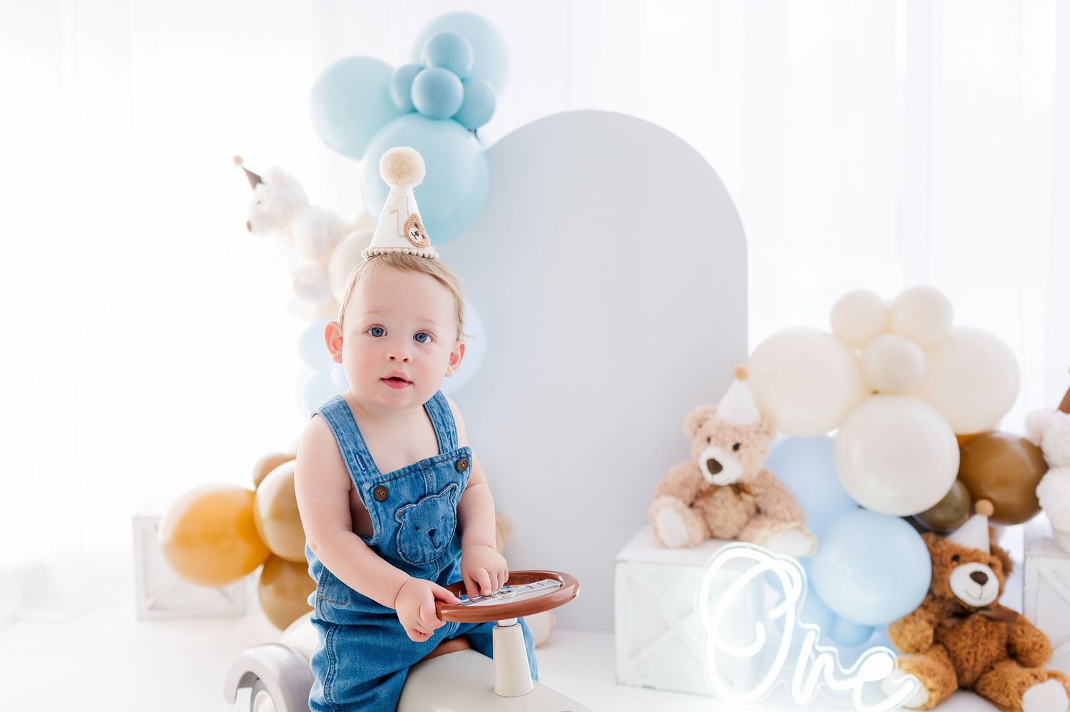 Baby in denim overalls sits by white arch with blue balloons and teddy bears at a first birthday party.