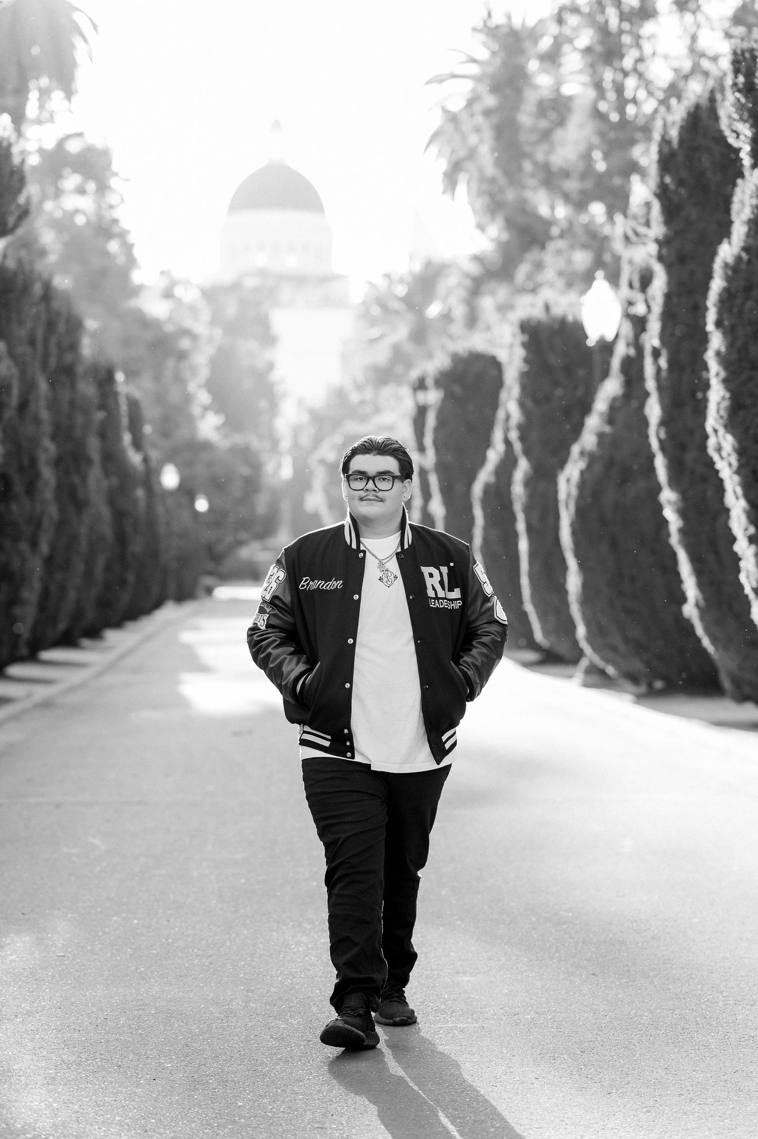 Black and white photo of a young man in a varsity jacket walking down a tree-lined path with a domed building behind him.