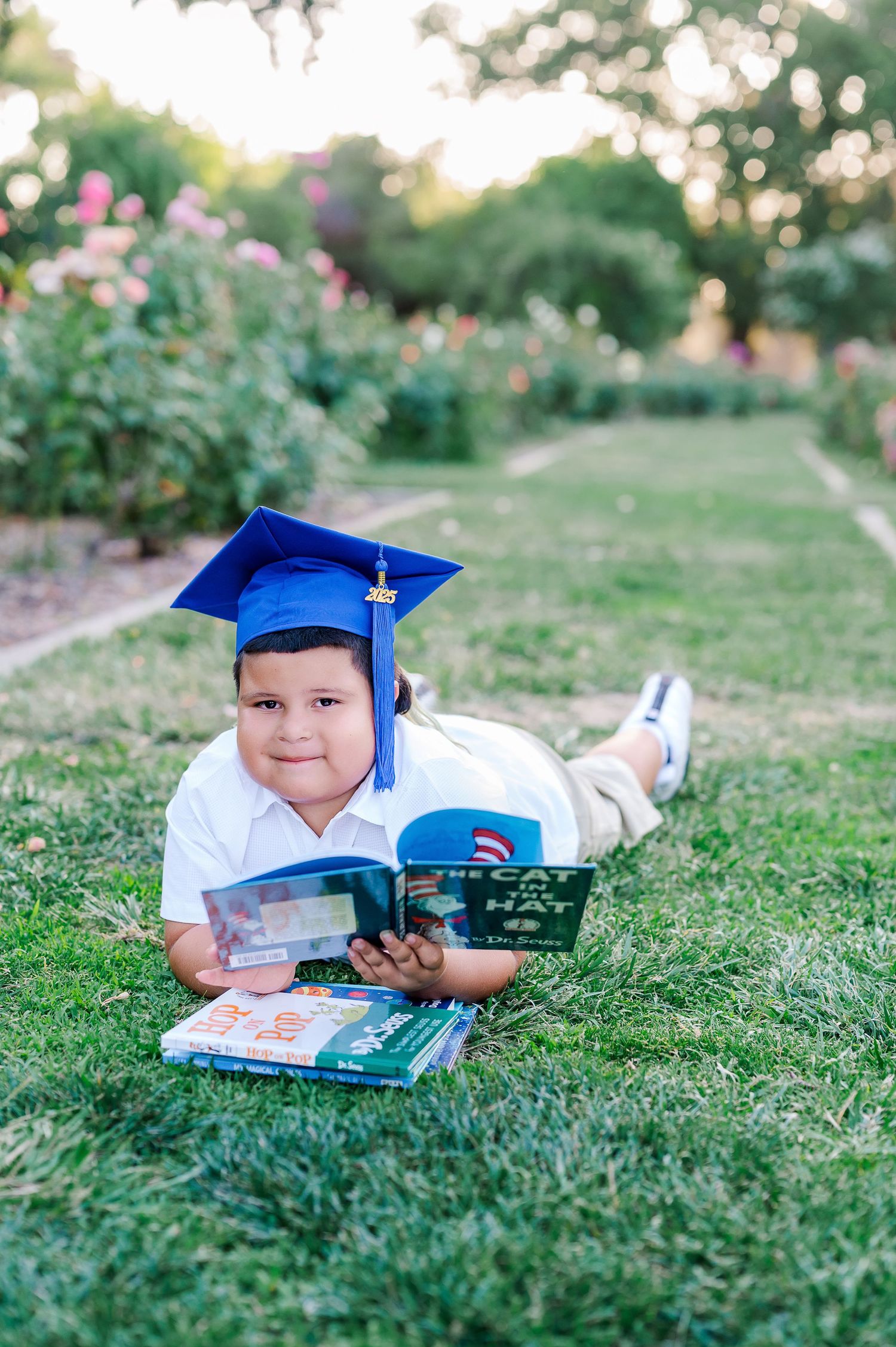 Young boy in blue graduation cap lying on grass reading books in a garden setting.