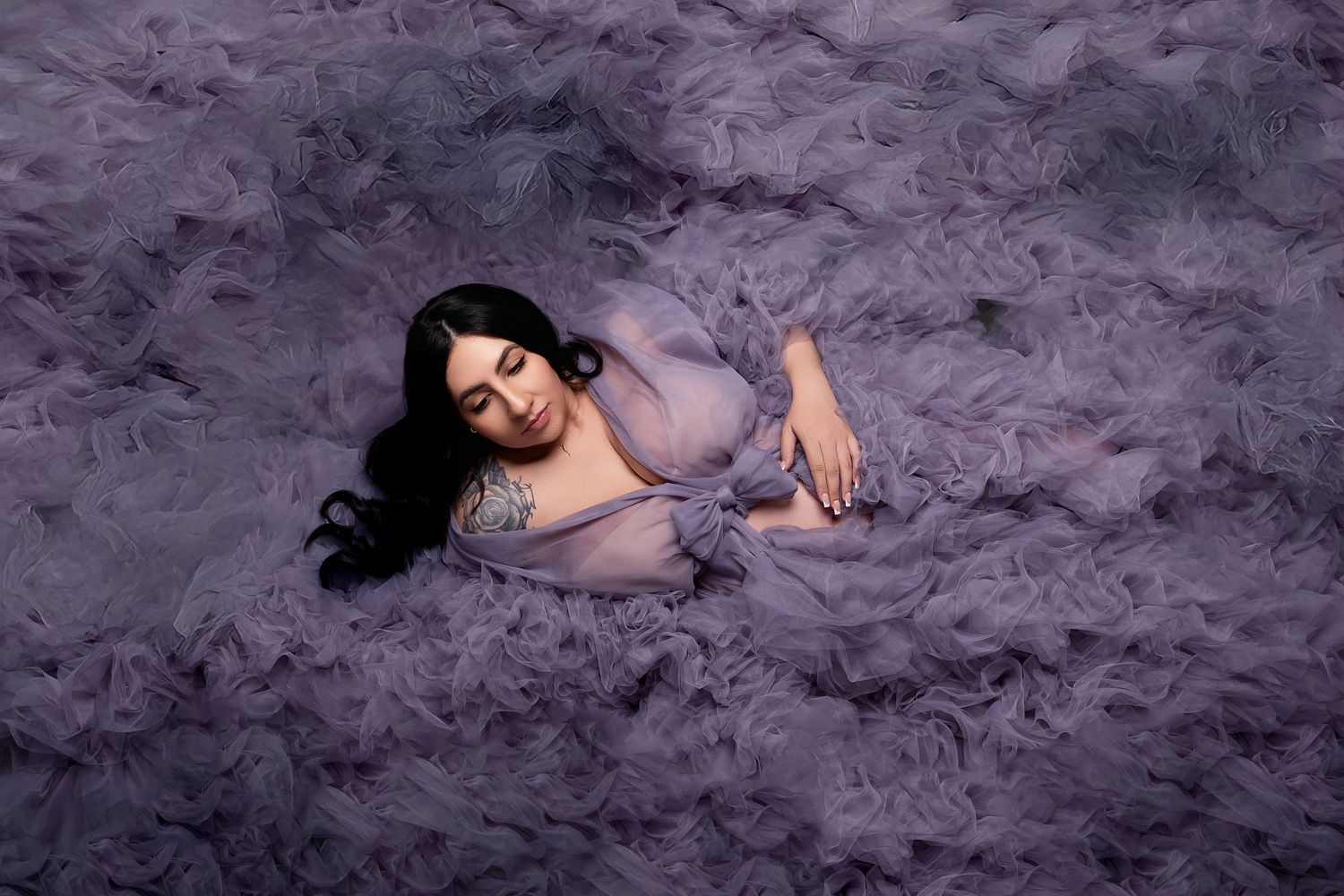 Artistic maternity photo with woman in purple dress lying on textured purple background.