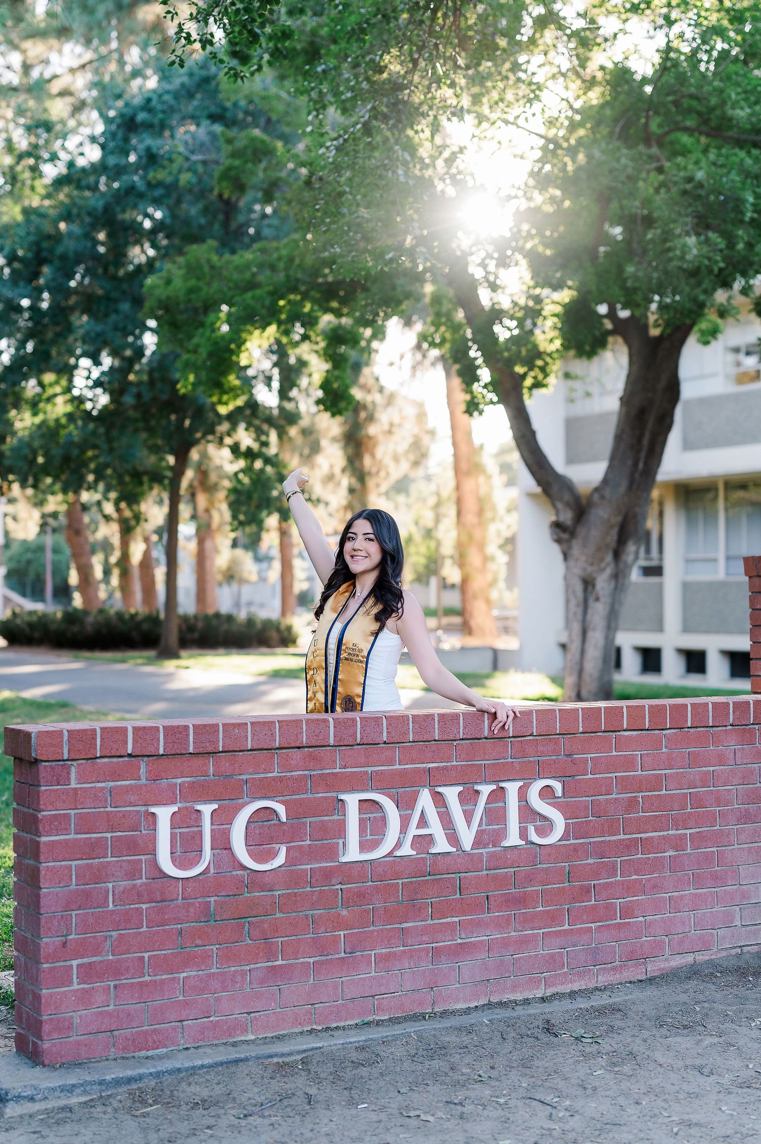 Graduate celebrating on the UC Davis brick wall with arms raised during sunset.