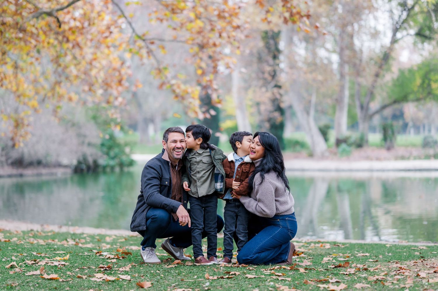 Family of four enjoying autumn day in park by pond, parents smiling at toddler children among fallen leaves.