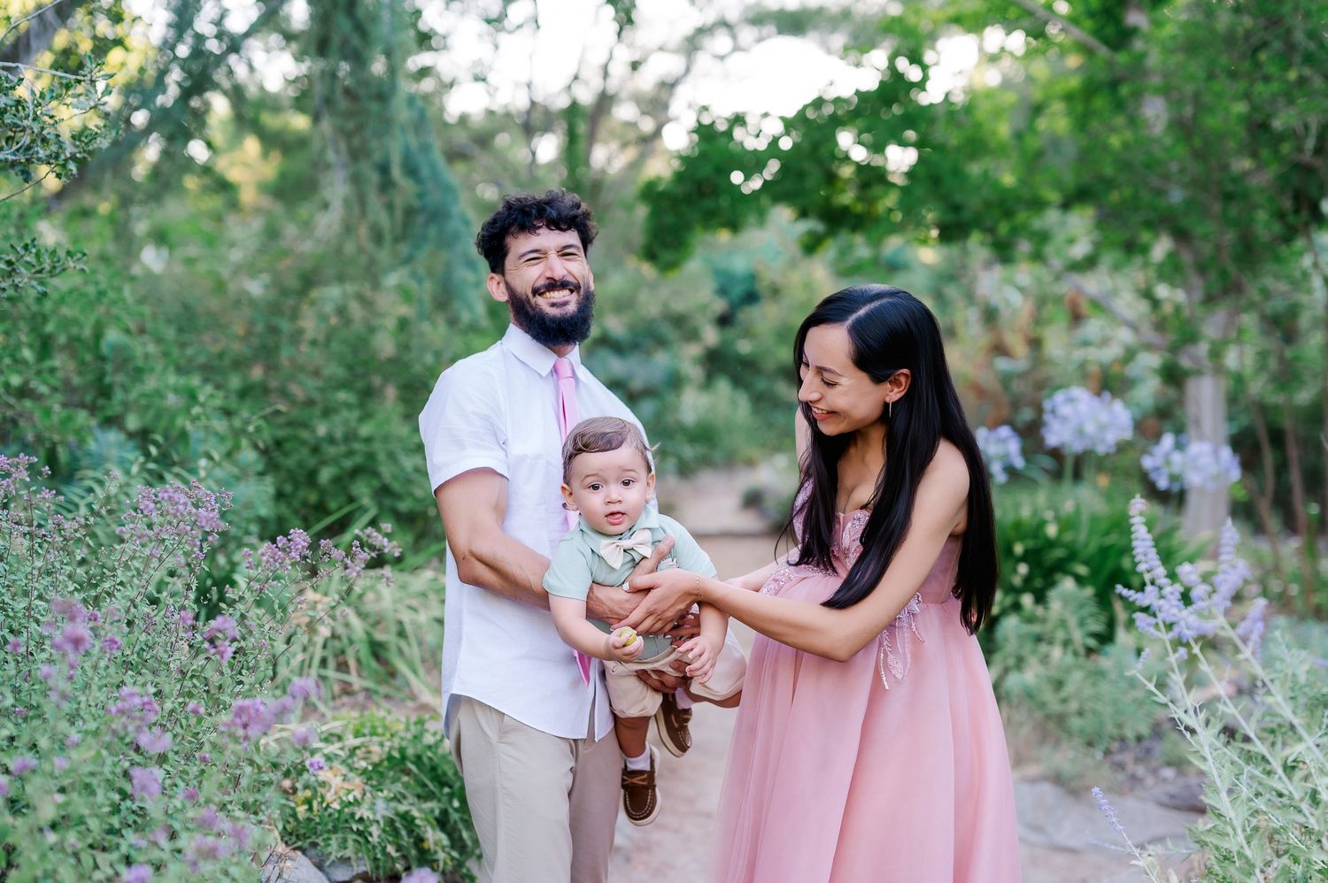 A smiling family with a bearded father, mother in pink dress, and baby enjoying a garden path surrounded by lush greenery. WPA Rock Garden