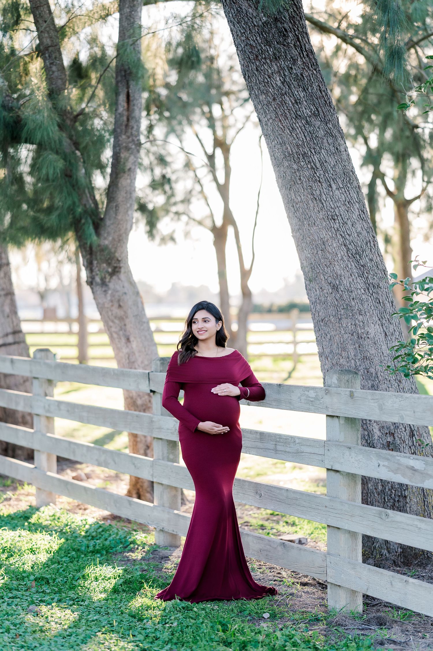 Maternity portraits along wooden fence with woman in burgundy dress in natural outdoor setting.