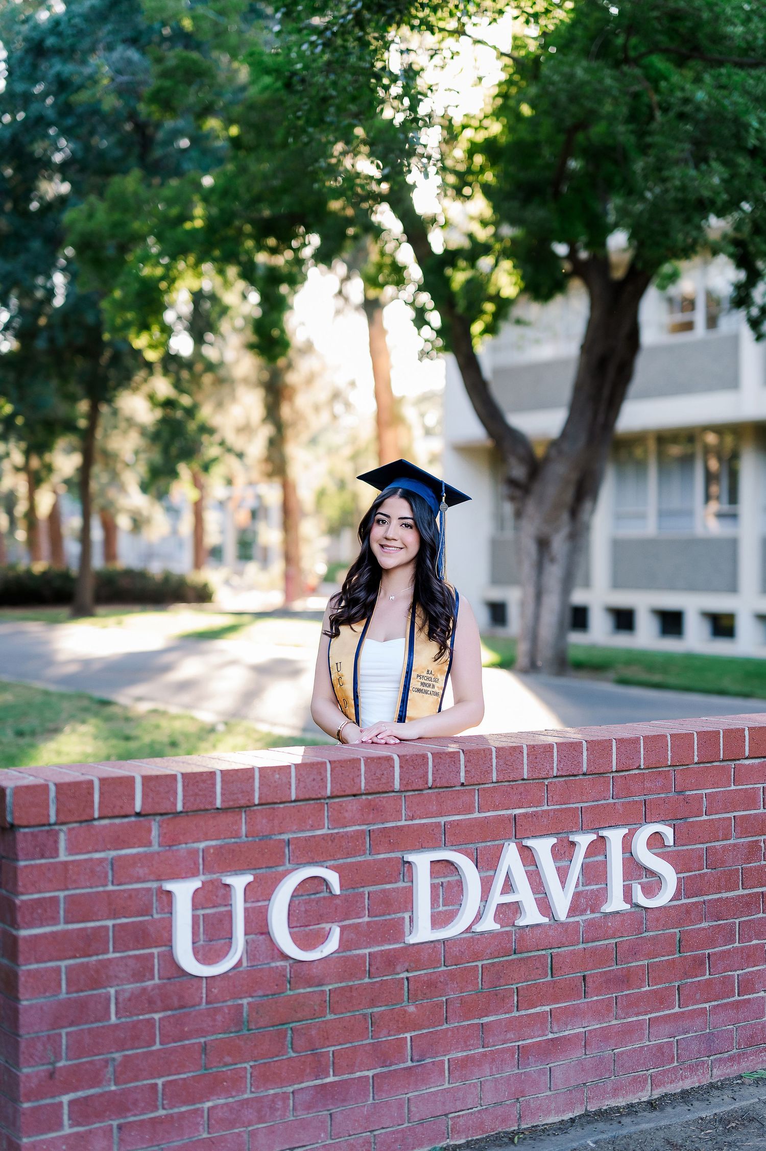 Graduate in cap and gown poses by UC Davis brick entrance sign during sunset.