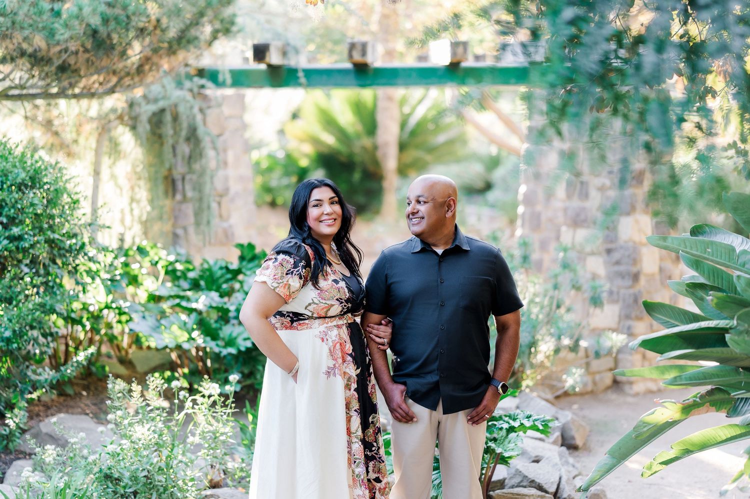 A couple shares romantic moments in a lush garden setting with stone walls and greenery in the background.
