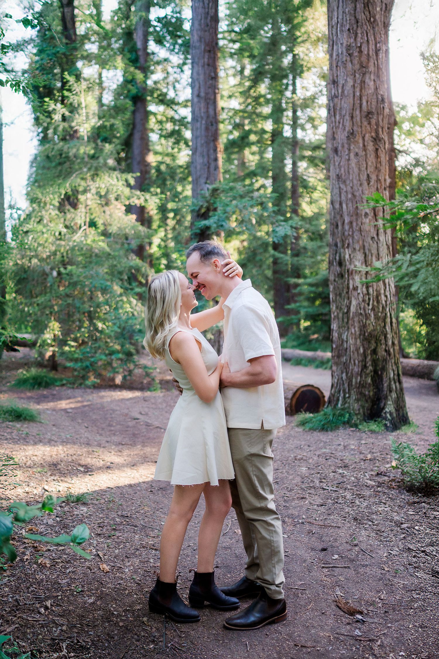 A couple shares an intimate moment on a dirt path surrounded by towering evergreen trees.