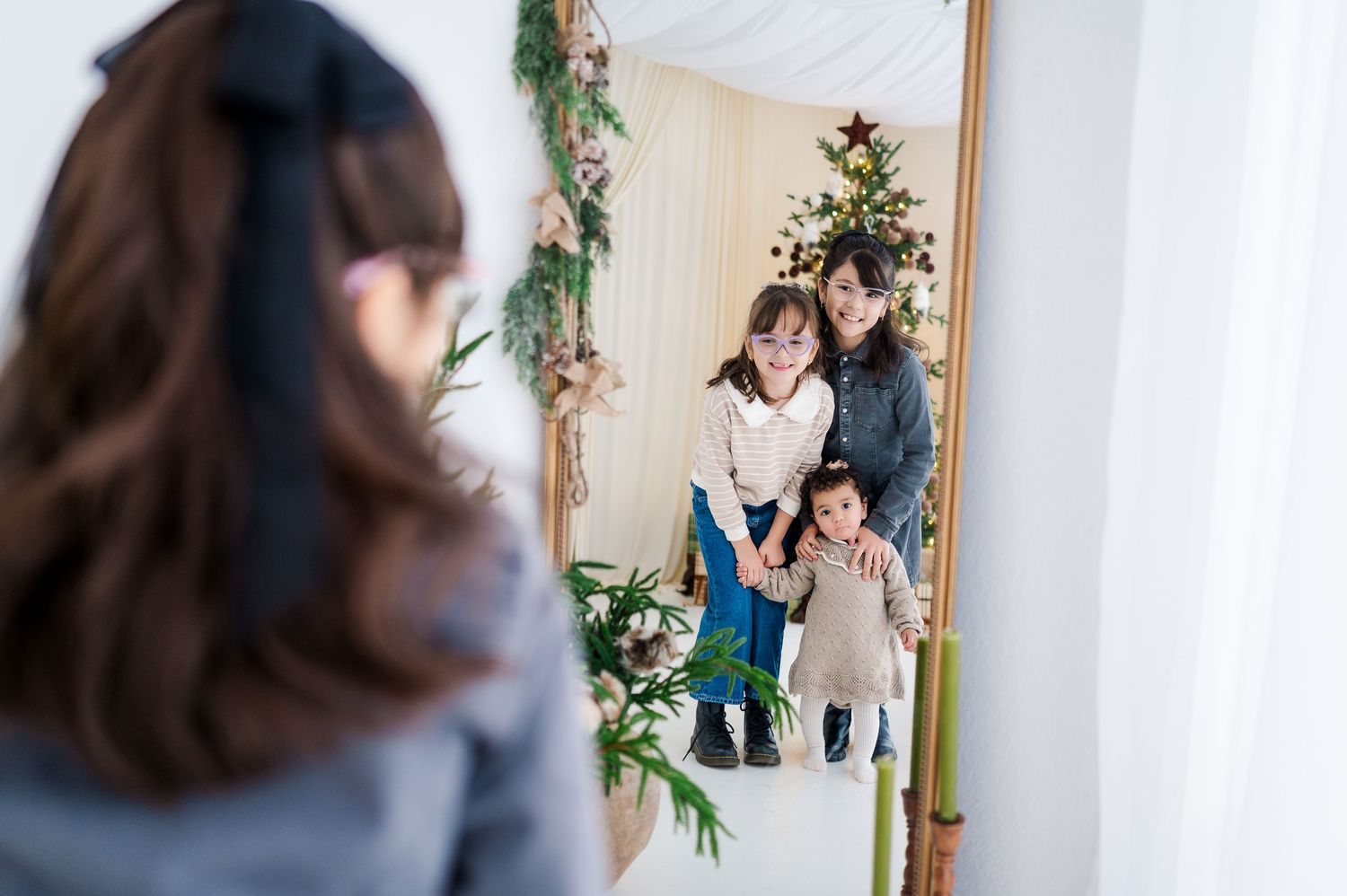 Family of three with a toddler smiling in a mirror reflection near a decorated Christmas tree.