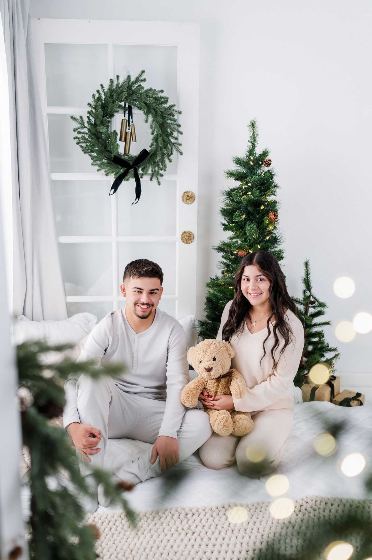 A couple in matching gray and white sweaters sits on a couch with teddy bears amid Christmas decorations.