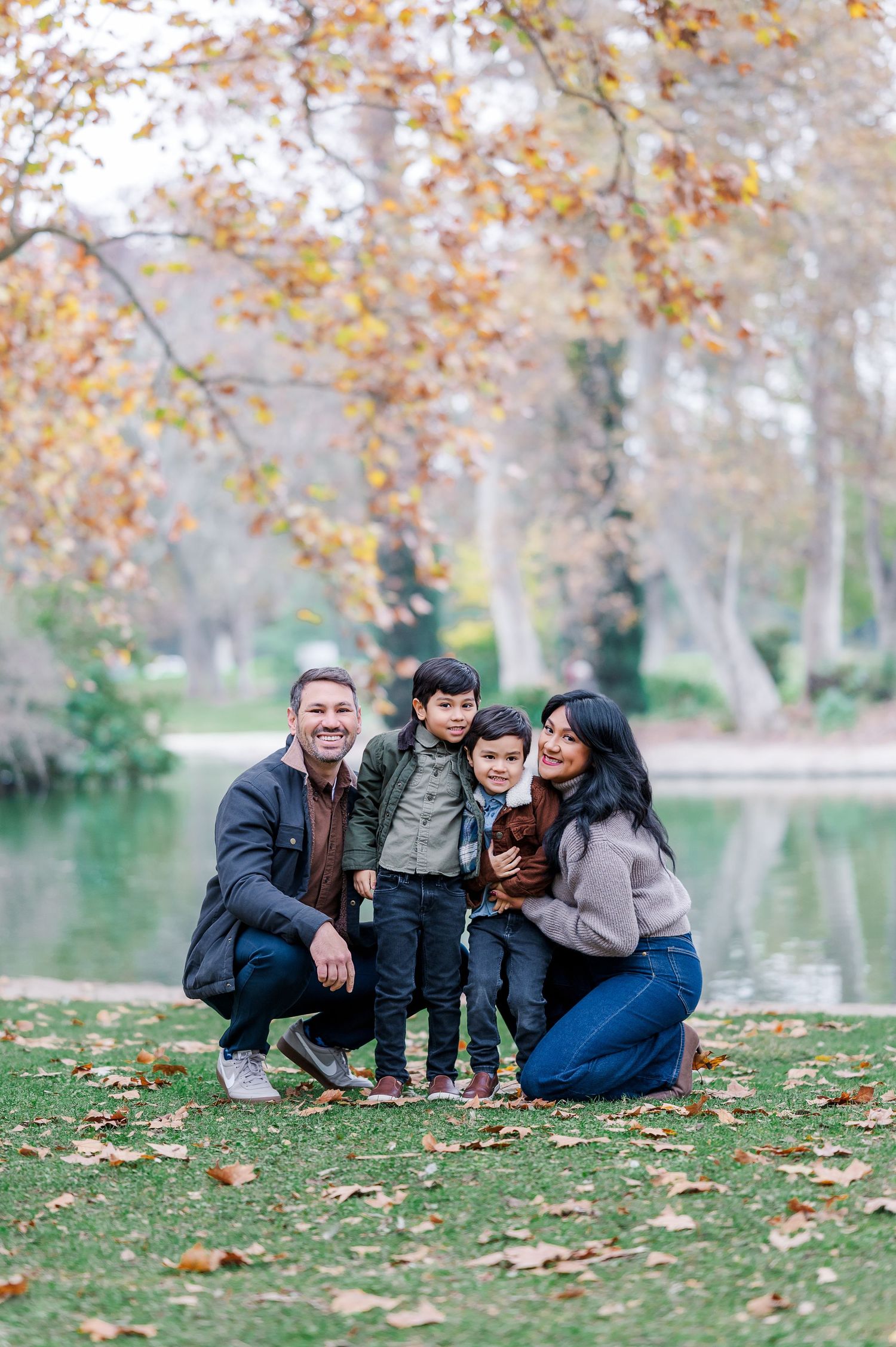 Family sitting together on autumn grass by lake with fallen leaves in park setting.