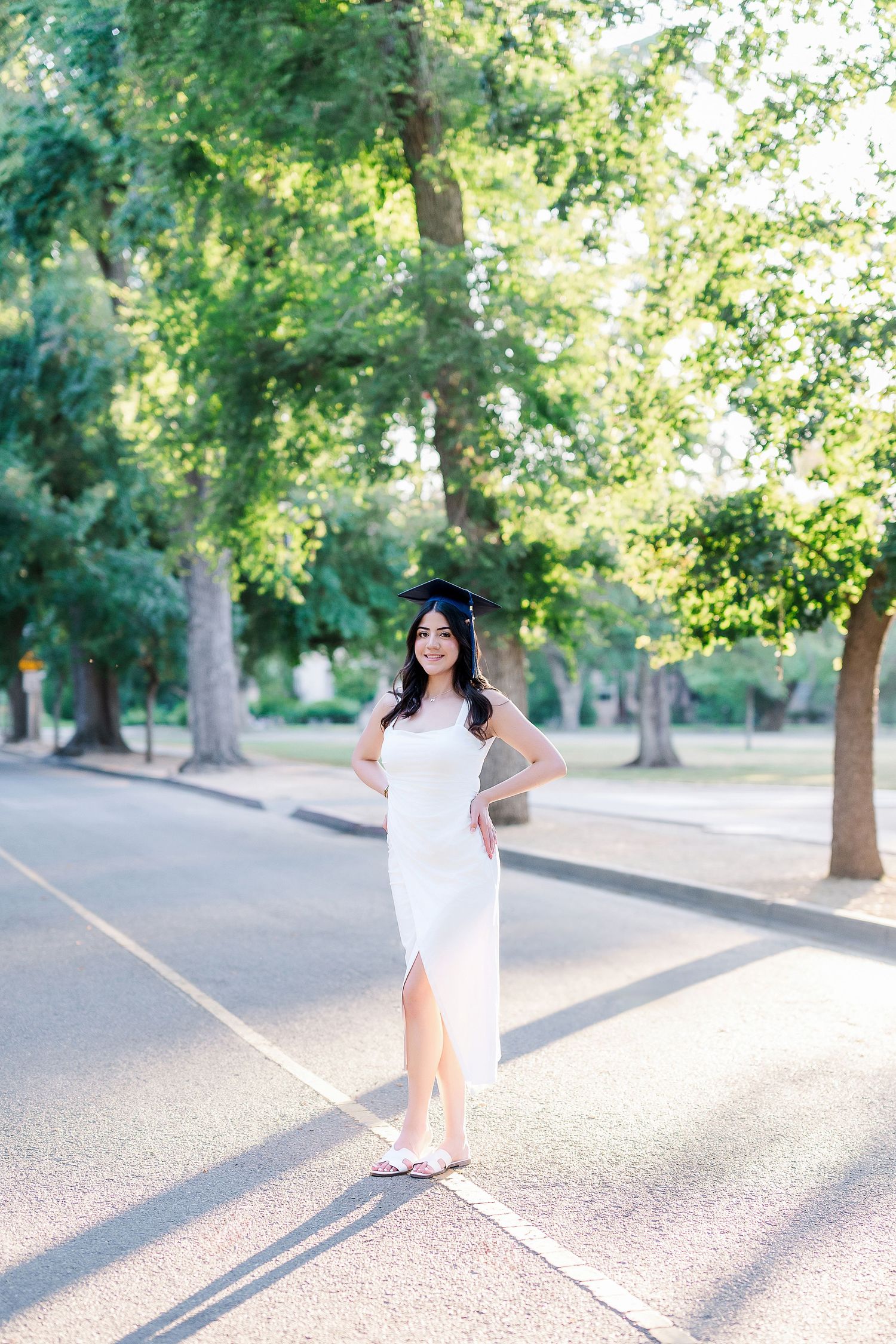 Graduate in white dress twirling and celebrating on campus pathway surrounded by trees.
