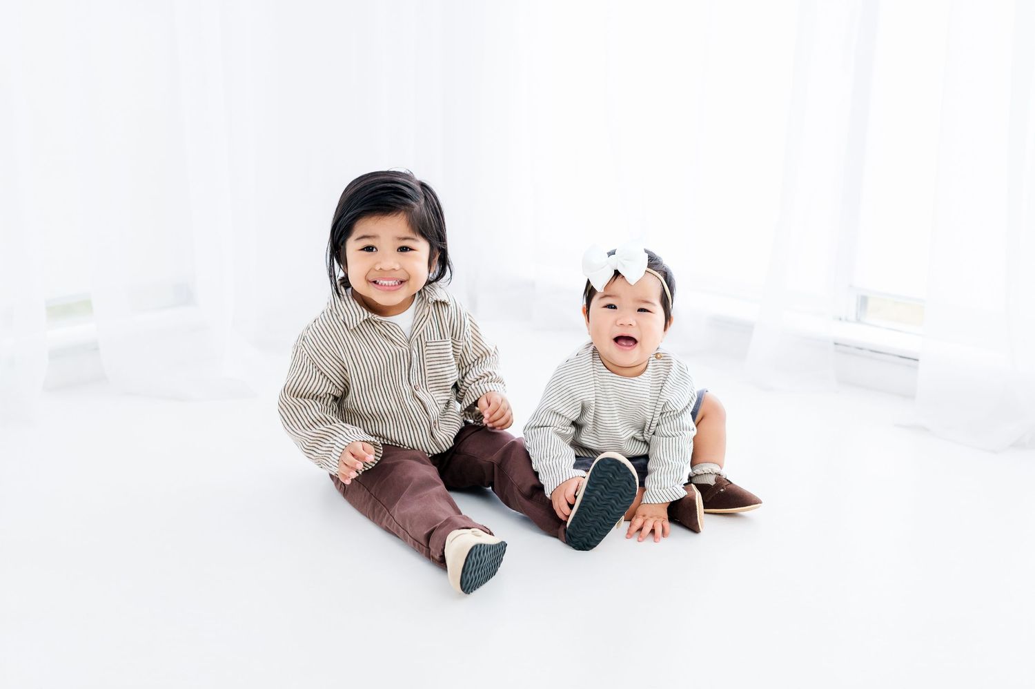 Two smiling Asian babies sitting together on white floor, wearing casual outfits, one with a bow headband.