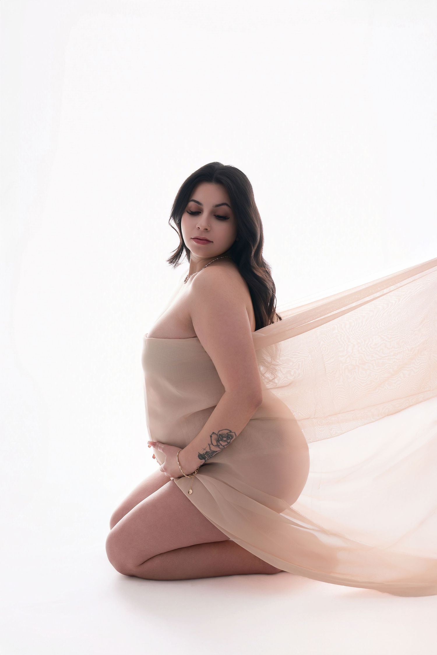 Pregnant woman in beige bodysuit with flowing fabric kneeling on white background in elegant maternity photo. Sacramento Maternity Photograp