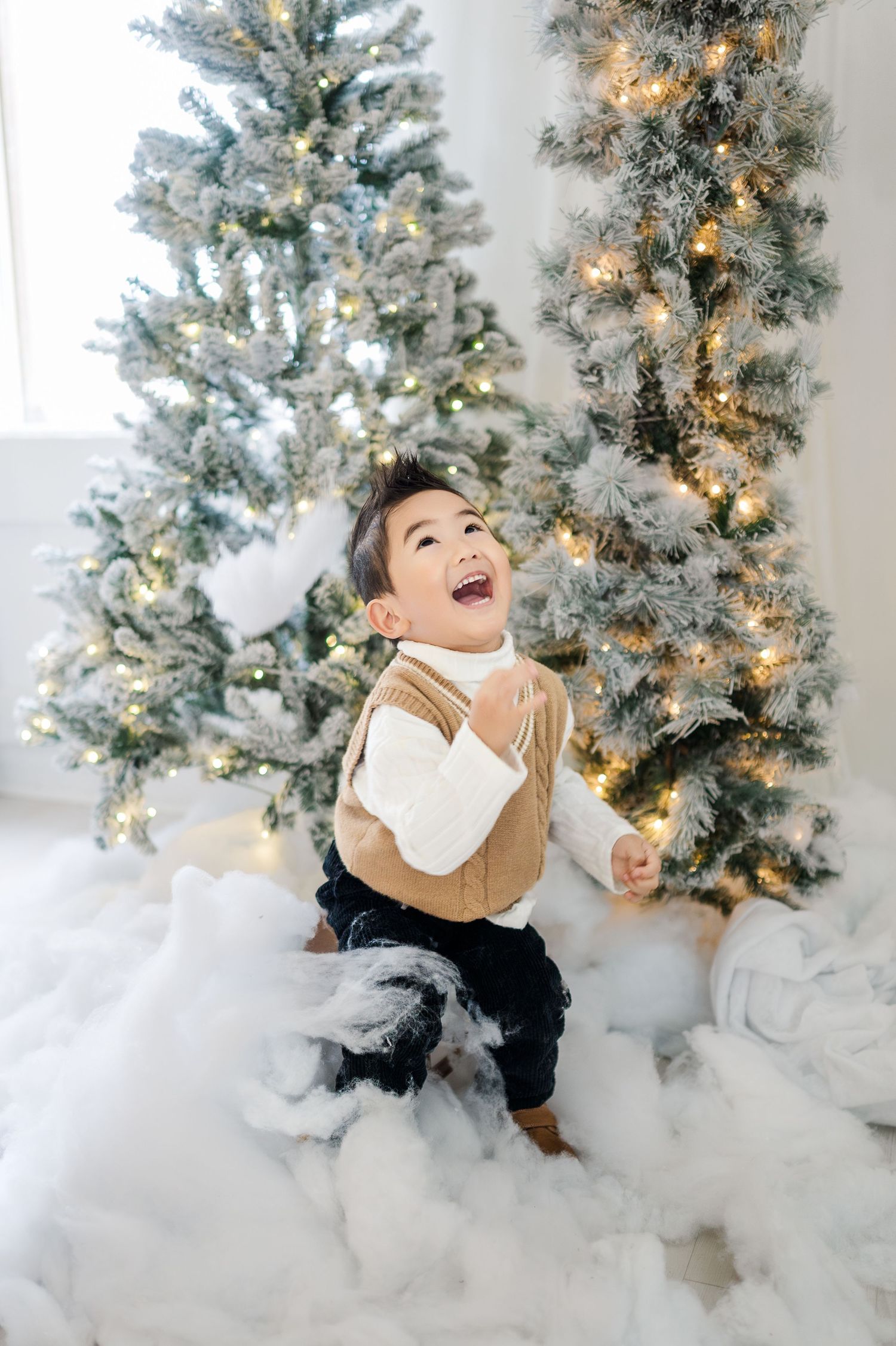 A small child in a brown vest laughs joyfully while standing in front of decorated Christmas trees and fake snow.