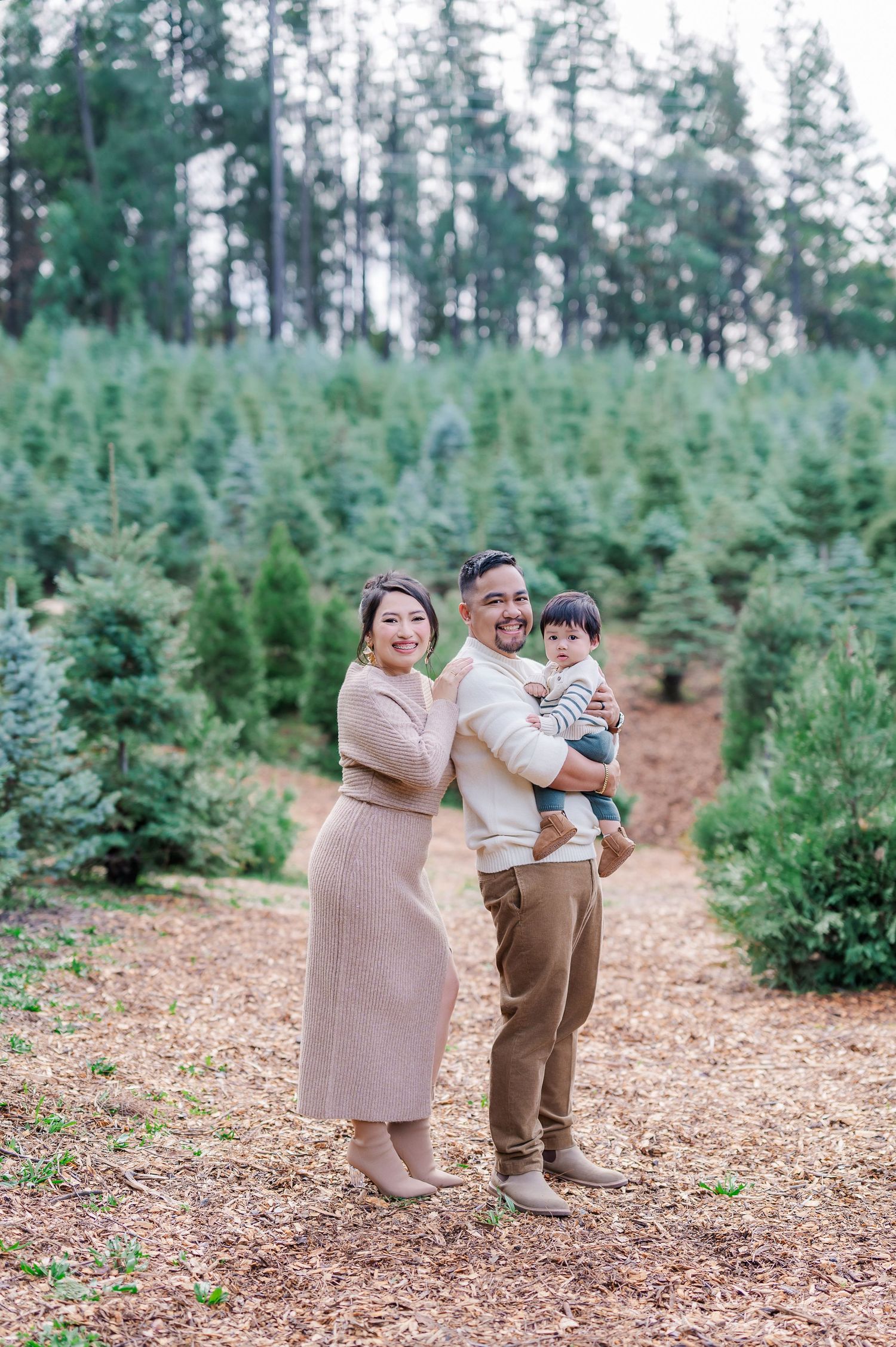Family poses together among evergreen trees at a Christmas tree farm on a sunny day.