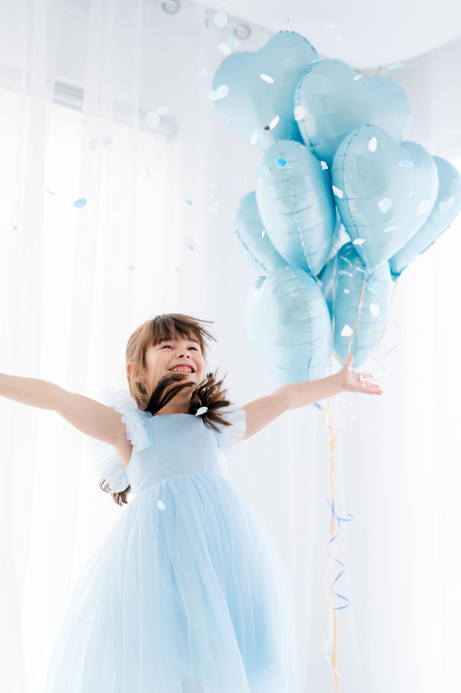 A joyful girl in a blue tulle dress holds heart-shaped blue balloons, surrounded by falling confetti in a bright room.