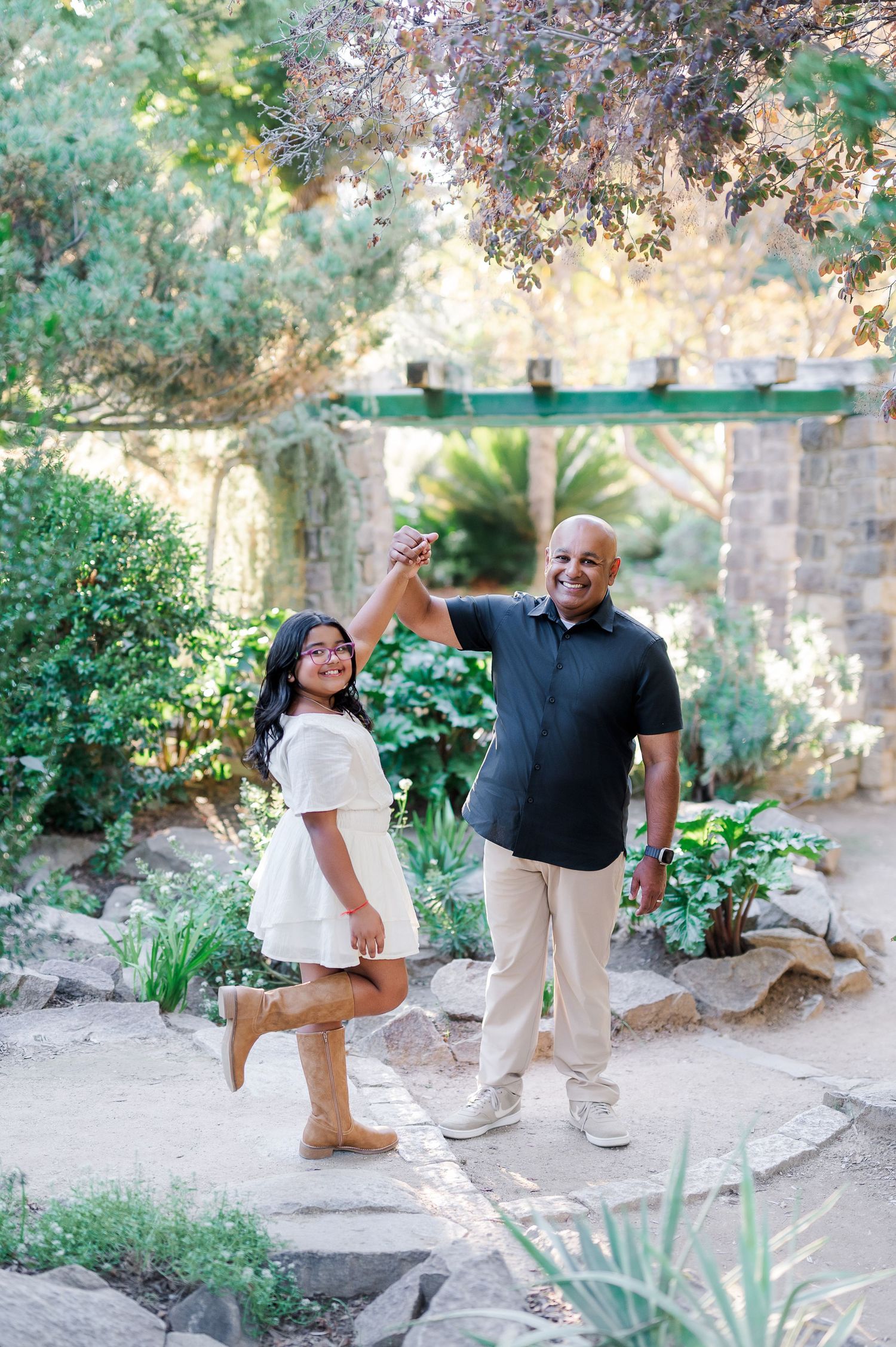 A couple poses together in a lush garden setting with stone walls and greenery during a sunny outdoor photoshoot.