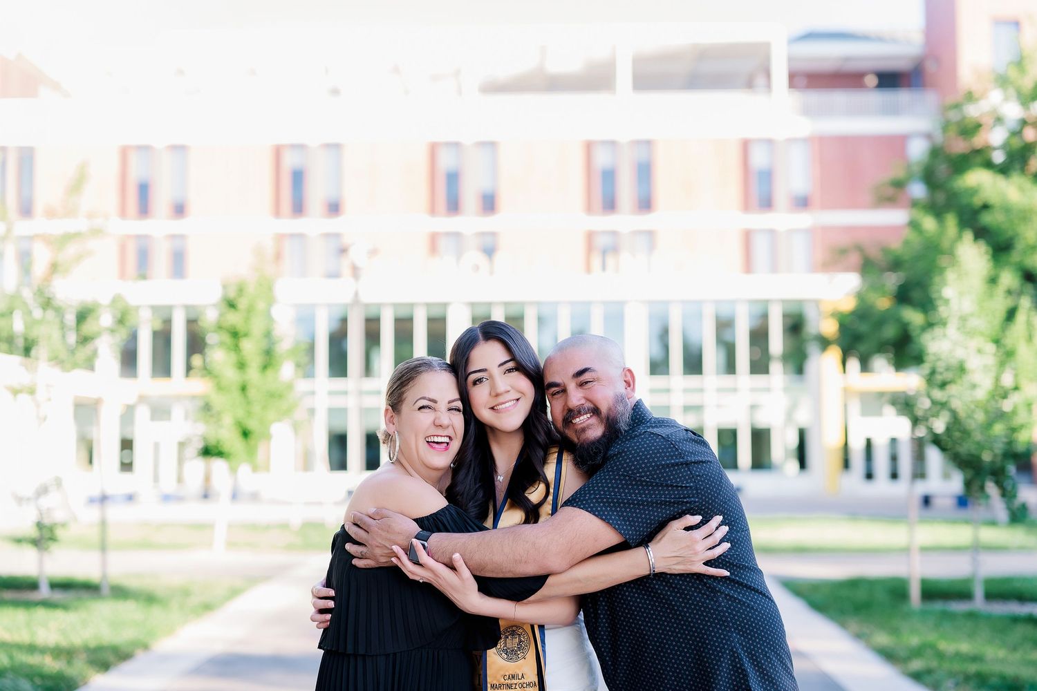 Graduation celebration with three people hugging on university campus lawn.