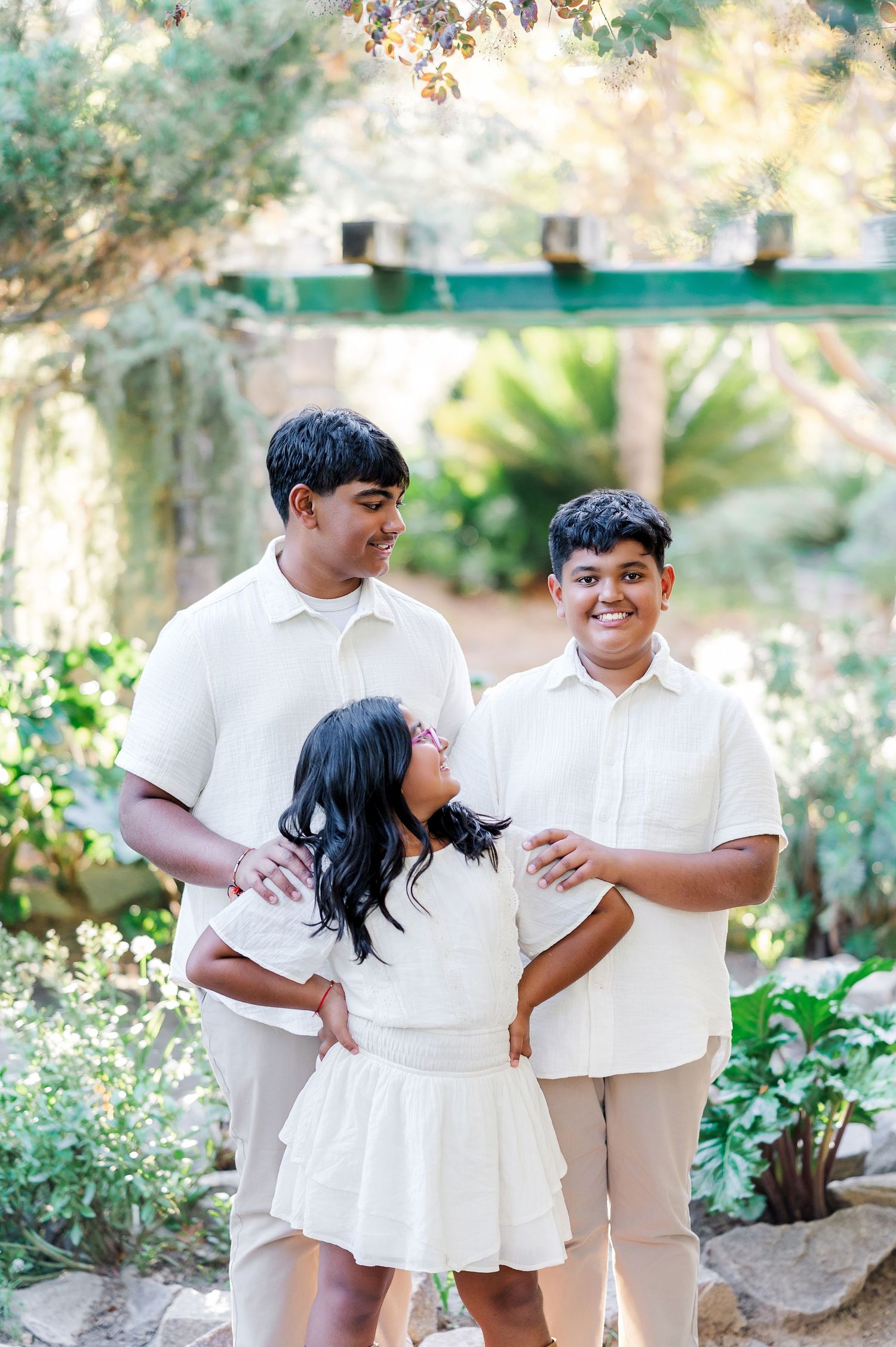 Three people in coordinated cream and white outfits share a joyful moment together in a sunny garden setting.
