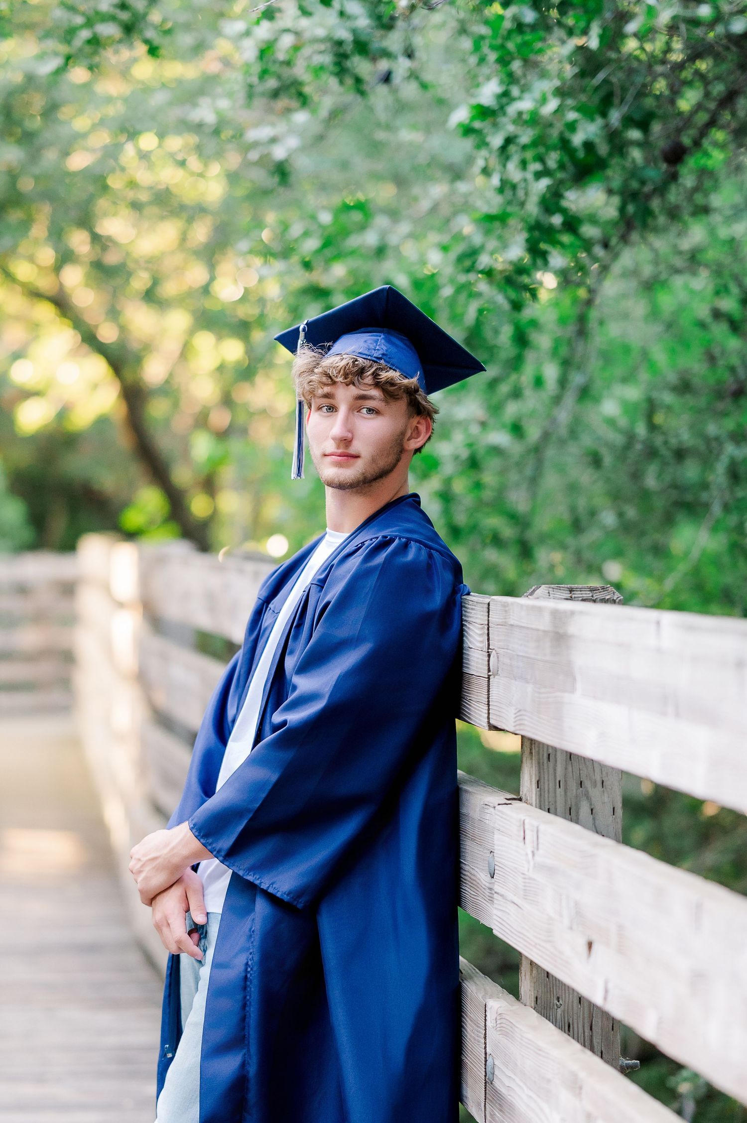 Young man in navy blue graduation cap and gown leaning against wooden bridge railing outdoors.