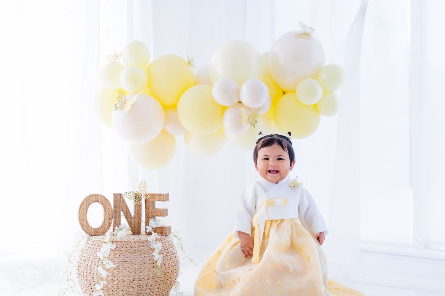 Baby girl in yellow dress celebrates first birthday with ONE sign and pastel balloon garland backdrop.